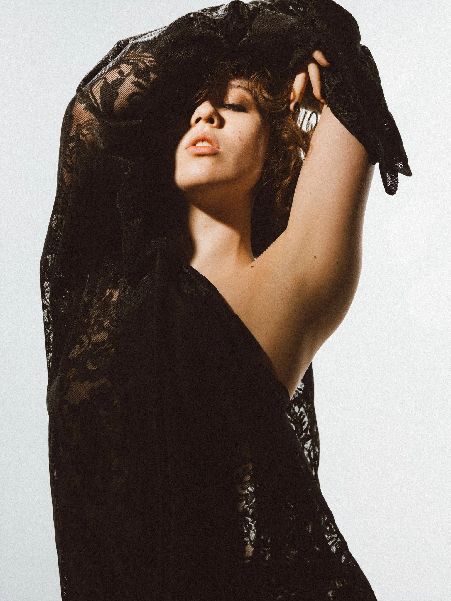 A woman with curly hair is raising her arm over her head, partially covering her face with a black lace garment. The photo is a close-up shot with a plain light gray background.