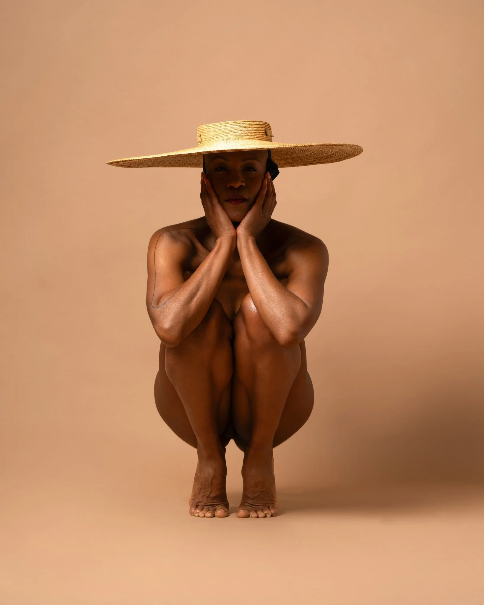 A person with a large sun hat covering their face, sitting cross-legged with hands on their cheeks, set against a plain peach background.