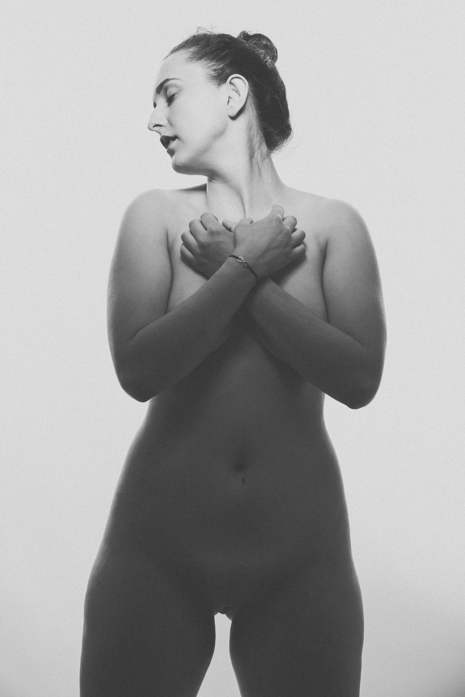 A black and white photo of a nude woman with her hair in a bun, her eyes closed, head turned to the side, arms crossed over her chest, holding her shoulders, standing against a plain background.