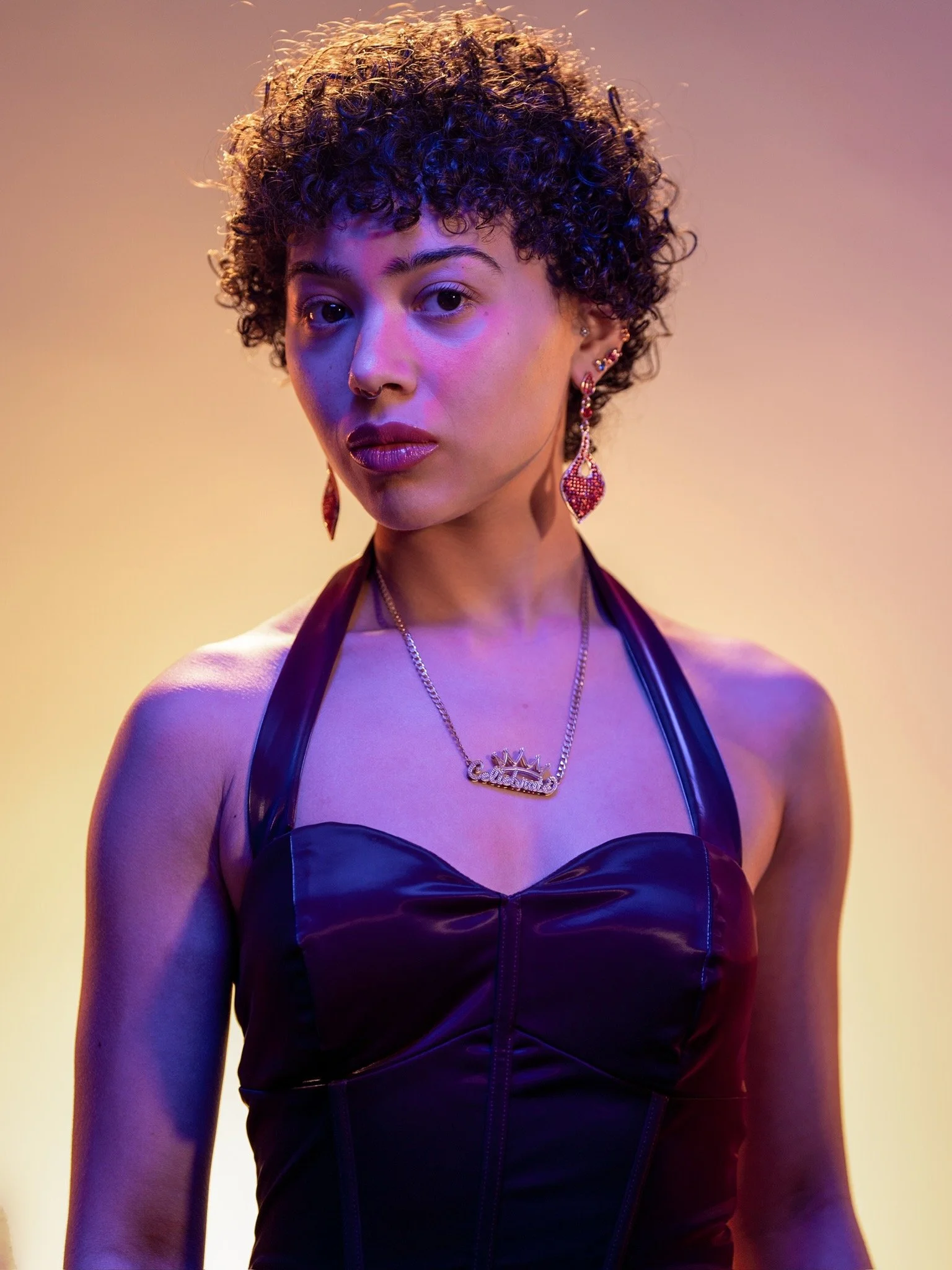 A woman with short curly hair, wearing colorful earrings, a necklace, and a black leather top, against a multicolored gradient background, illuminated with purple and yellow lighting.
