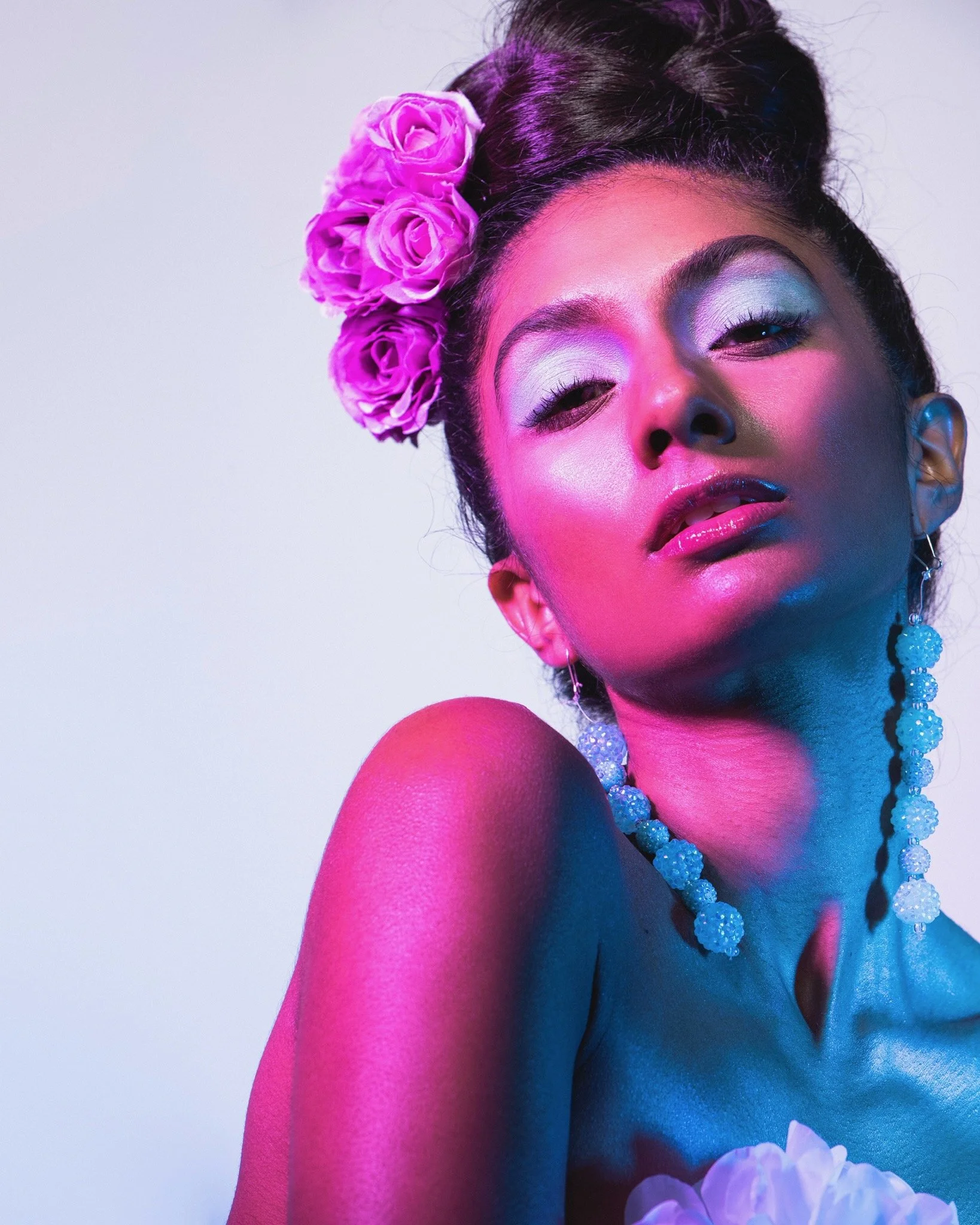 Close-up of a woman with dark hair wearing pink and purple makeup, large pearl earrings, a beaded necklace, and pink roses in her hair, against a plain background.