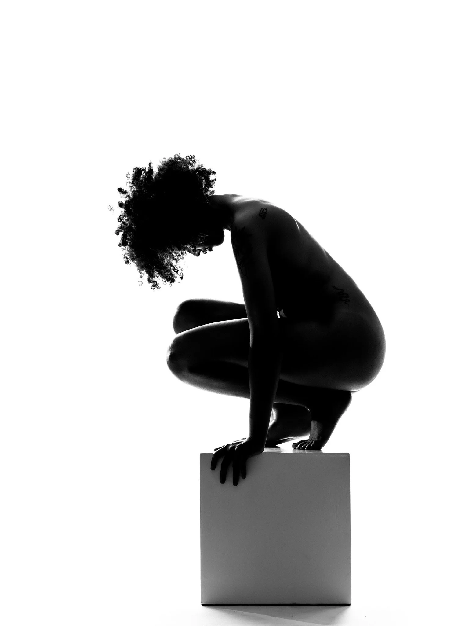 Silhouette of a woman with curly hair crouching on a pedestal, surrounded by a bright white background.