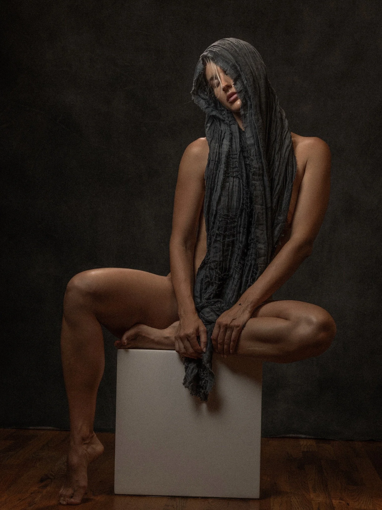 A woman with a black and gray hooded scarf covering her head and face, sitting on a white block, with her arms and legs positioned in a artistic pose against a dark background.