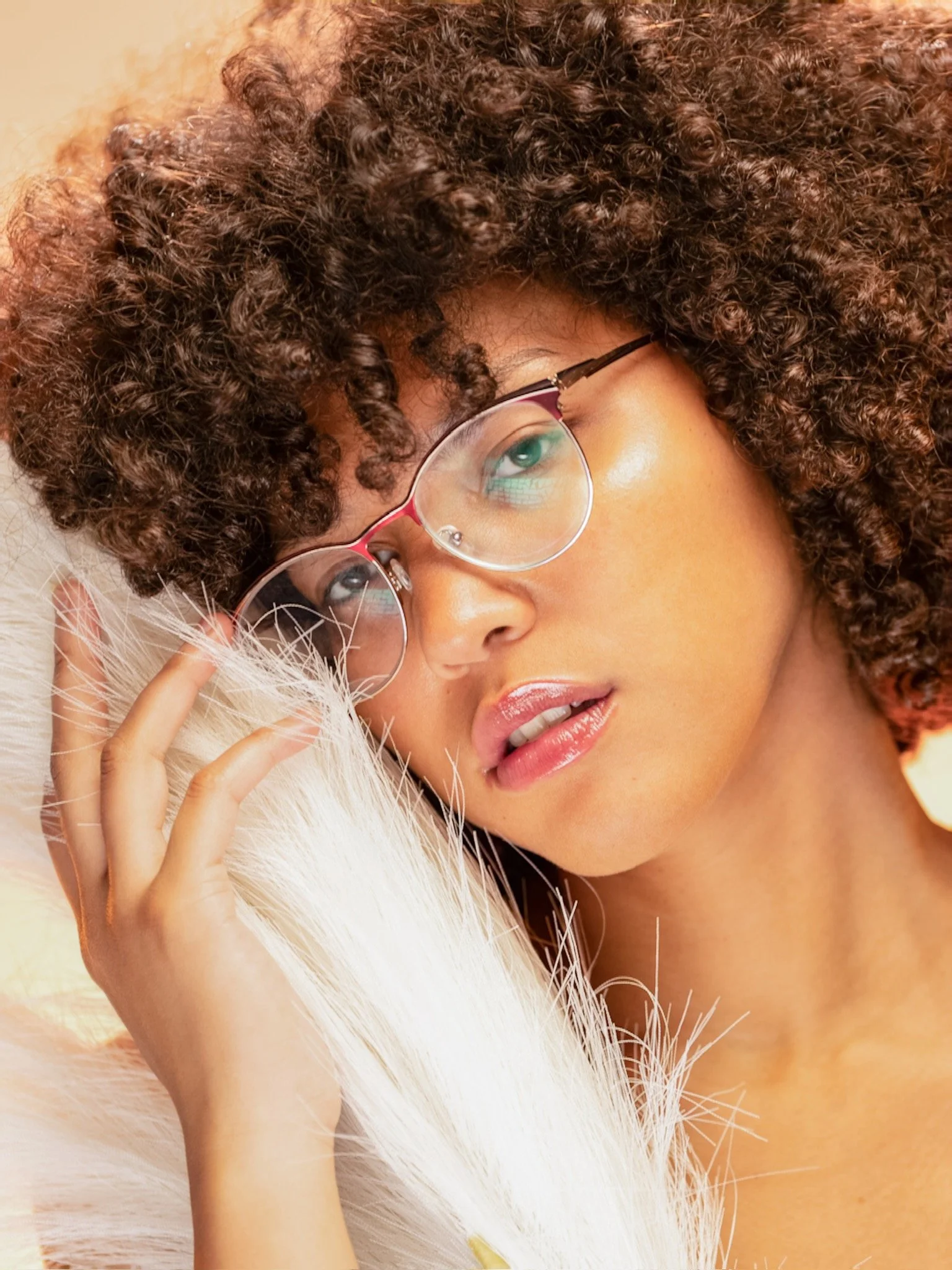 Close-up of a woman with curly brown hair, wearing glasses, lying on a white, feathery surface, with soft lighting highlighting her face and hair.