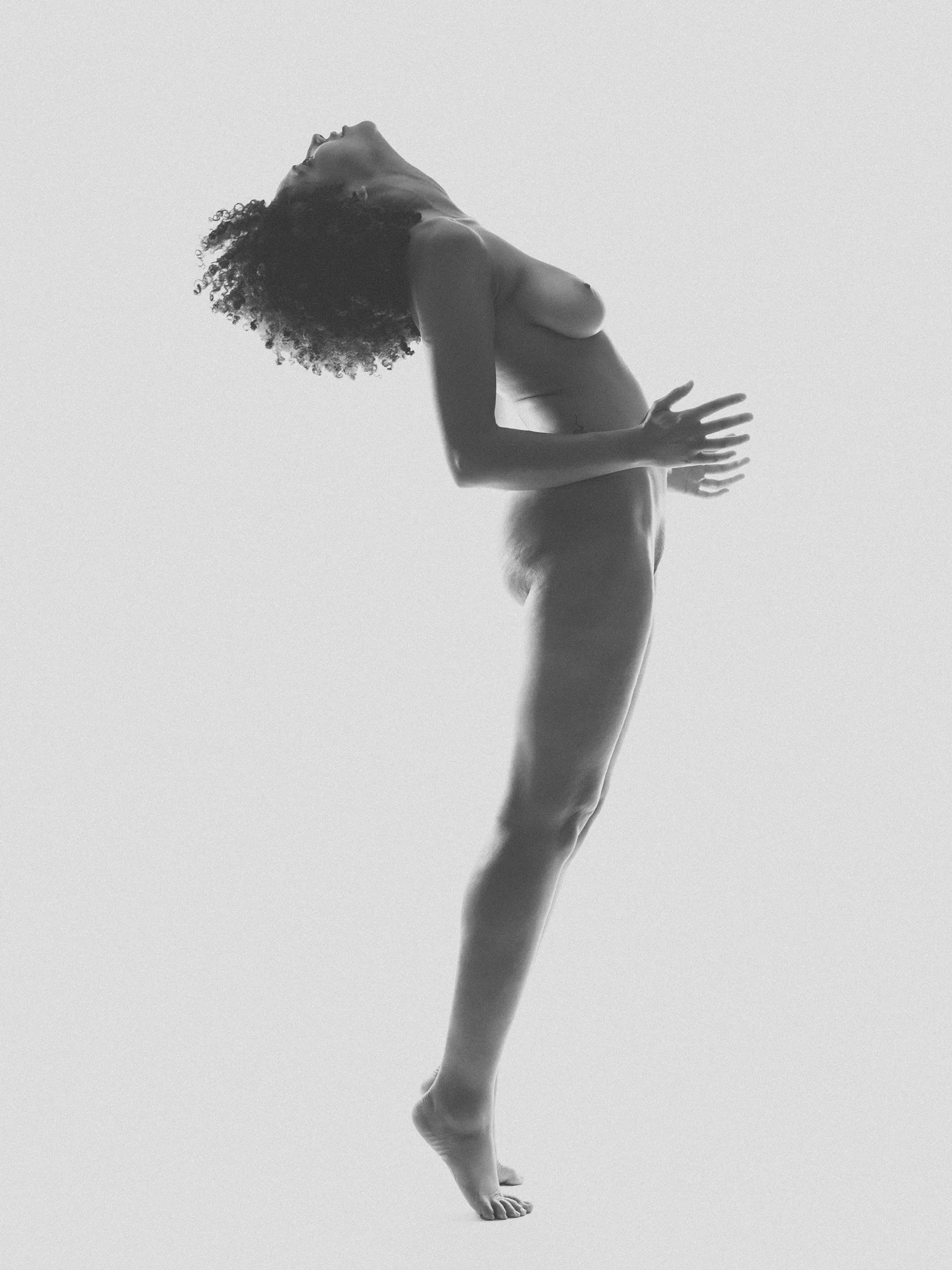 A black and white photo of a naked woman performing a headstand with her head and hands on the floor, her body extended vertically, and her curly hair hanging downward.