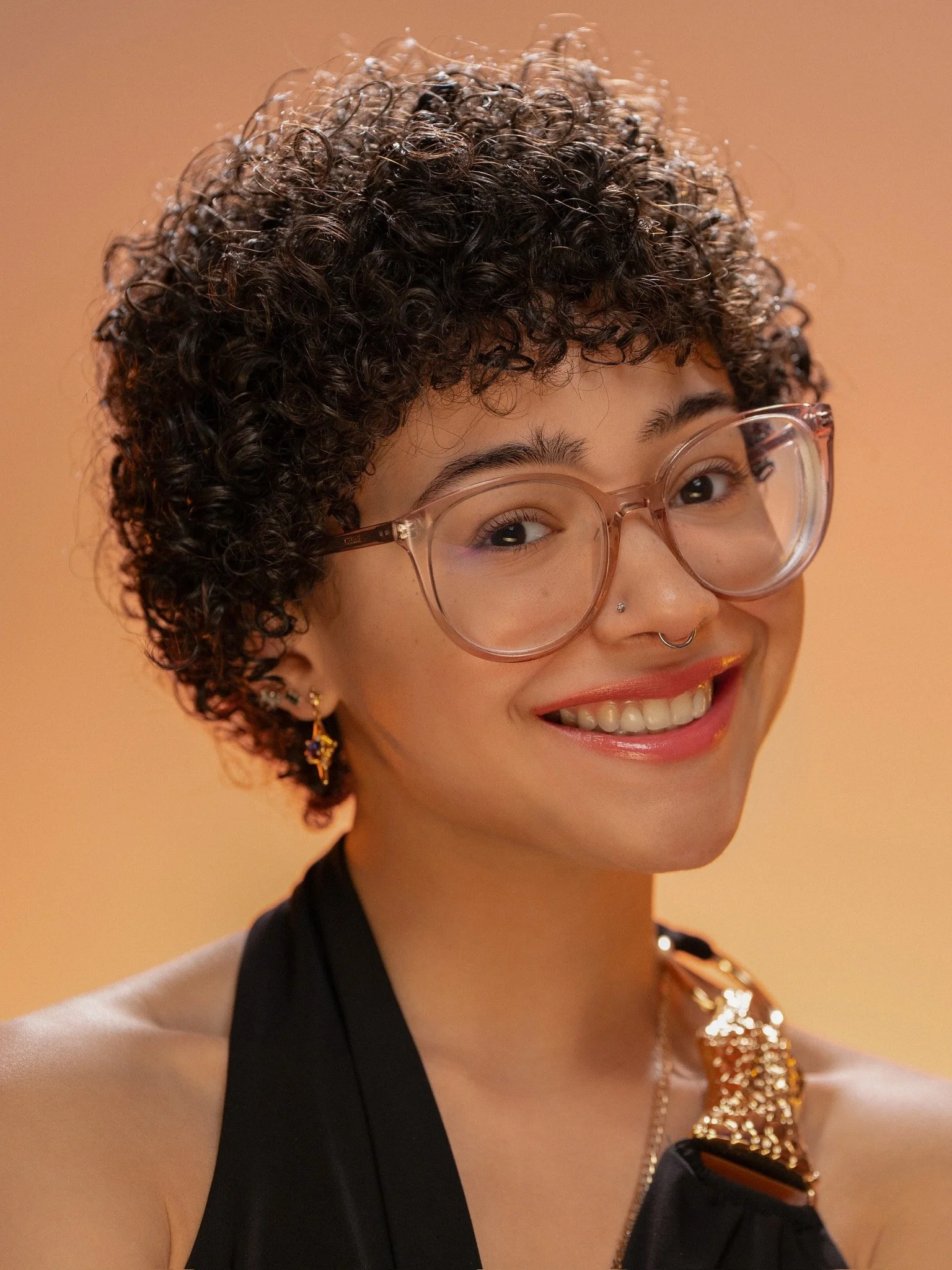 A woman with curly short hair, wearing glasses, earrings, and a nose piercing, smiling, dressed in black and white clothing, against a beige background.