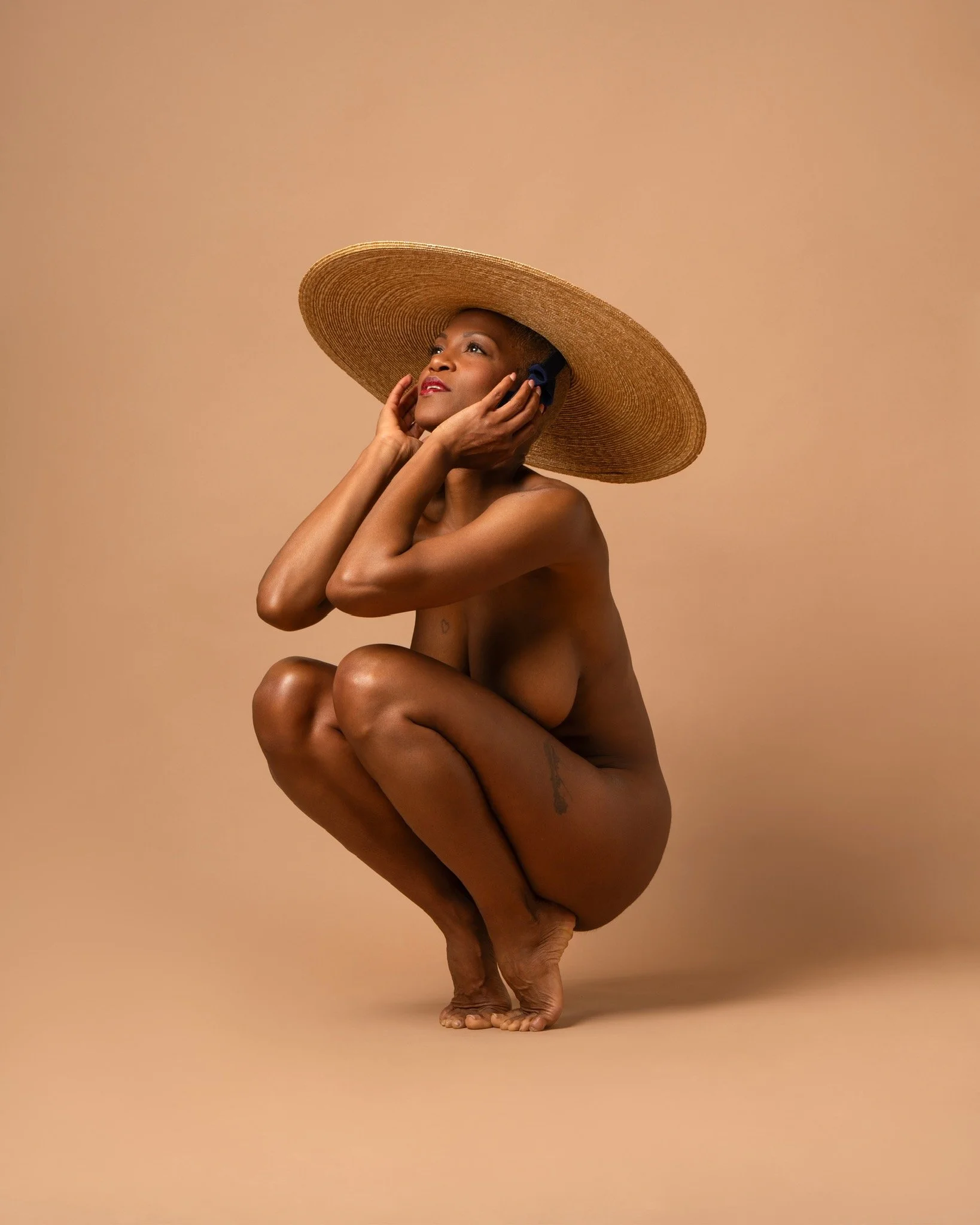 A person with dark skin crouches on the ground, wearing a wide-brimmed straw hat and headphones, with their hands adjusting the headphones on their head, against a solid beige background.