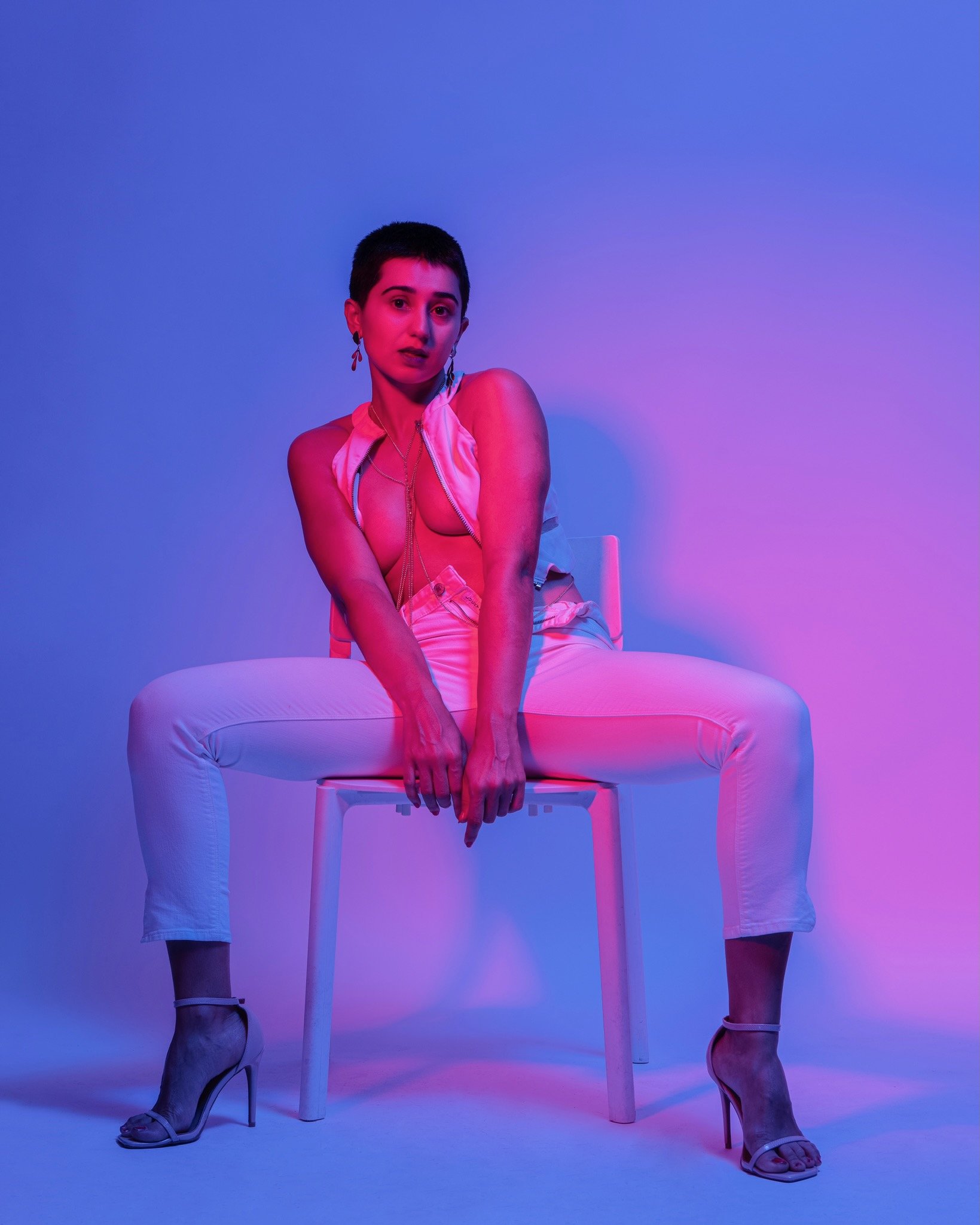 A woman with short dark hair and light skin tone sits on a white chair with her arms crossed, wearing a sleeveless satin top, white pants, and high-heeled sandals, illuminated by pink and purple lighting against a plain background.