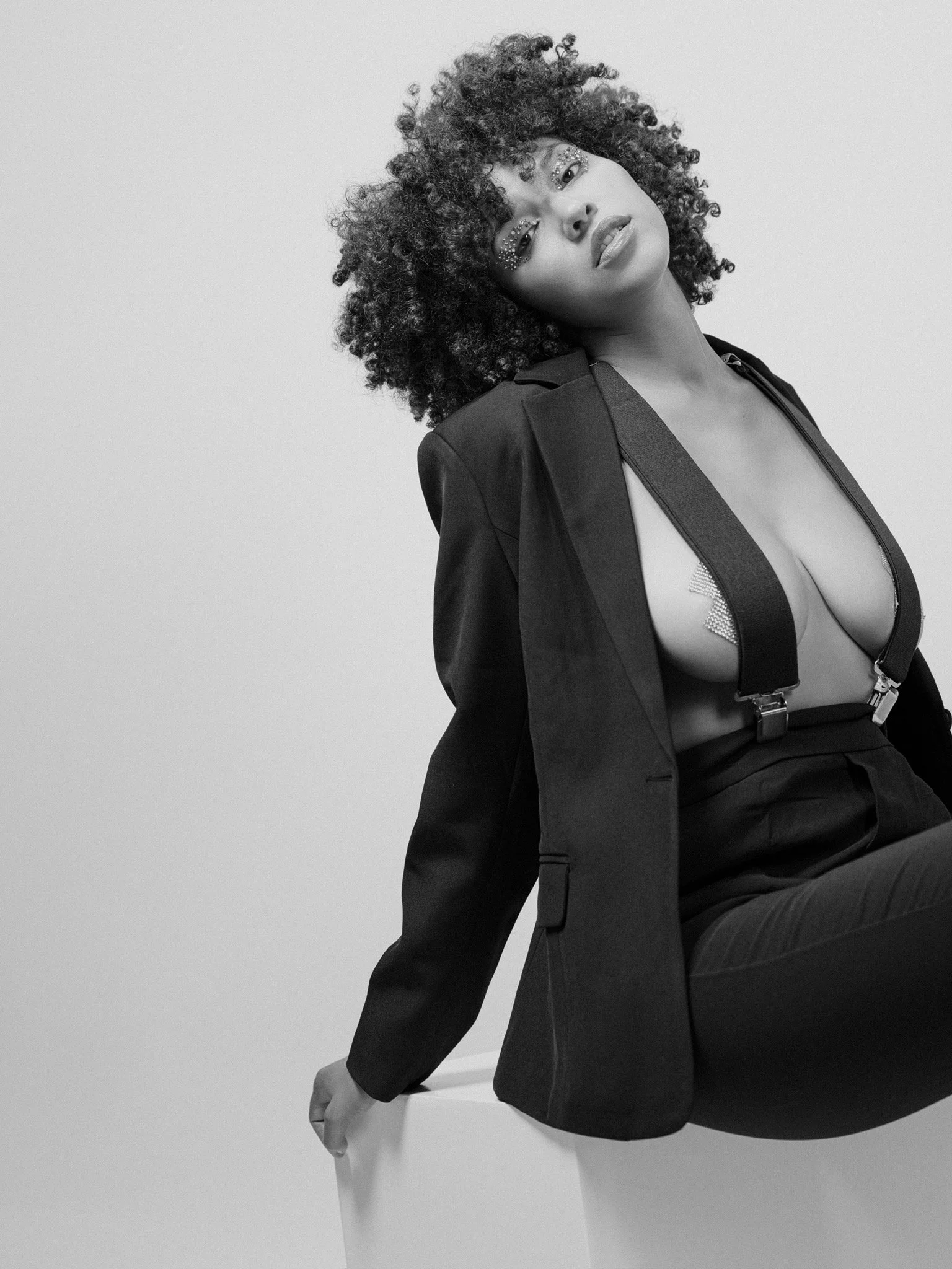 Black and white photo of a woman with curly hair, wearing makeup with embellishments around her eyes, dressed in a blazer with suspenders, posing against a plain background.