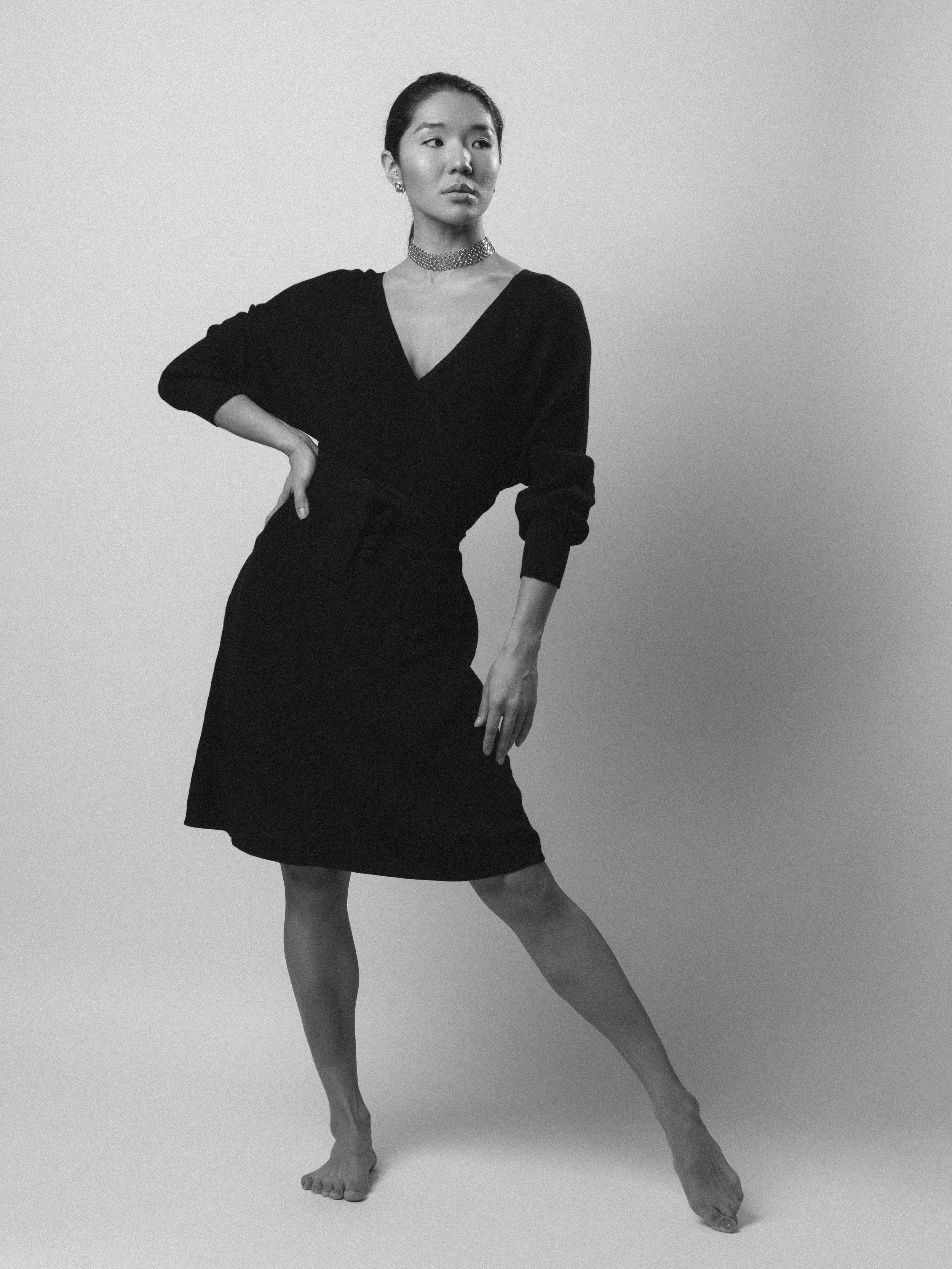 A woman in a black dress, wearing jewelry, posing with one hand on her waist and the other hanging down, against a plain background.