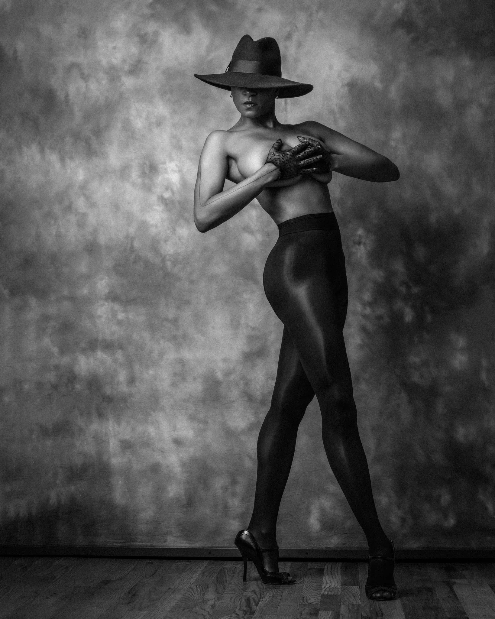 Black and white photograph of a woman in high heels, wearing a wide-brimmed hat covering part of her face, black gloves, and a dark skirt, posing against a textured background.