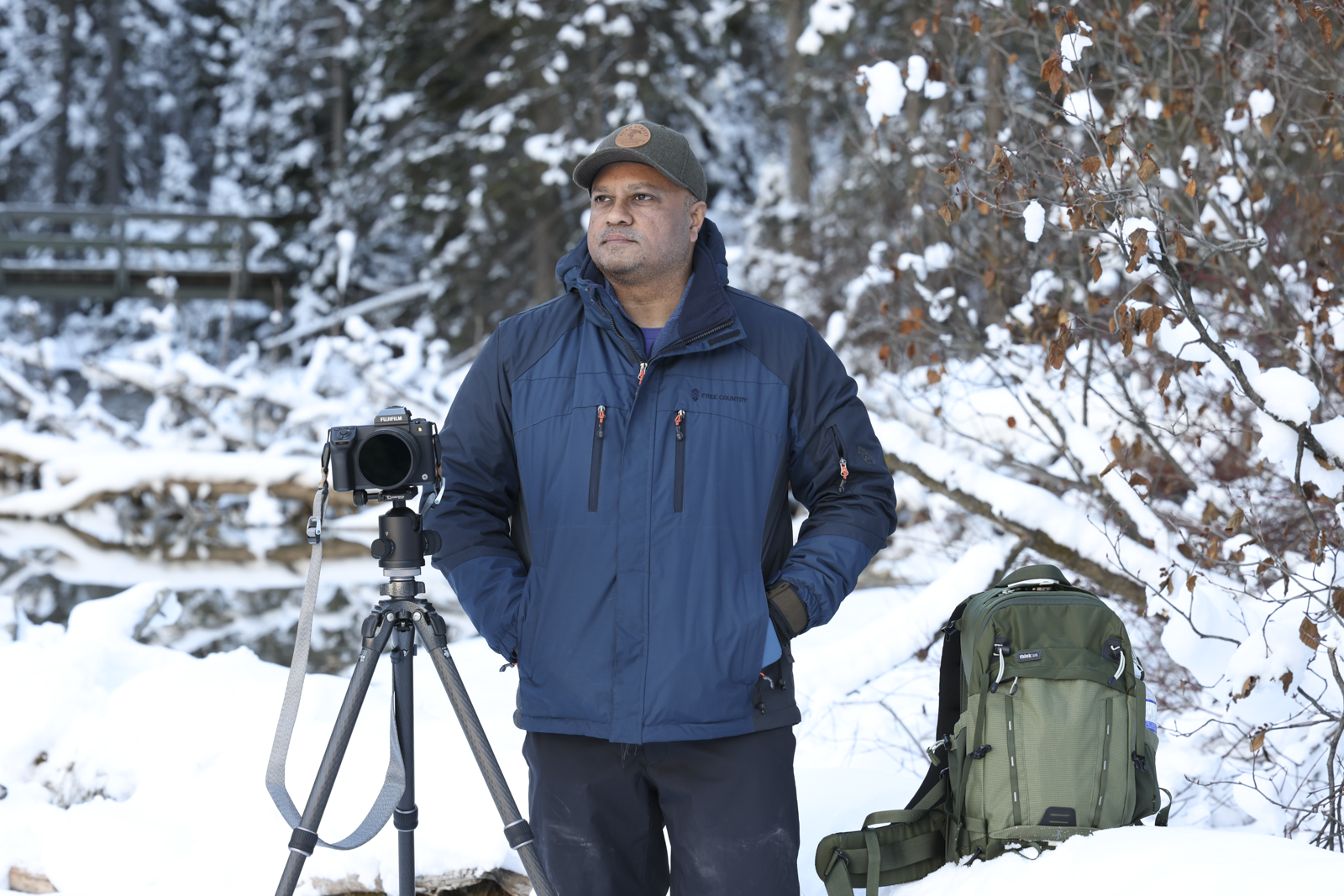 A man standing in snowy woods with a camera on a tripod, wearing a blue jacket and a gray cap, with a green backpack on the snow beside him.