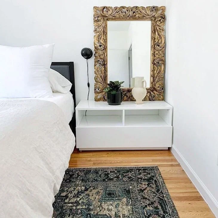 A bedroom corner with a white nightstand, a large ornate gold mirror, a potted plant, and a white vase on the nightstand. Part of a bed with white bedding and pillows is visible on the left. A navy patterned rug and wooden floor complete the scene.
