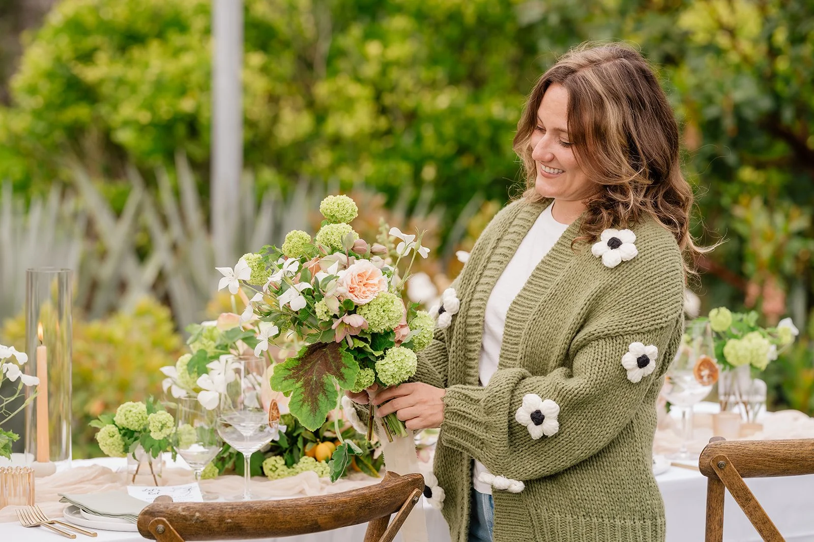 Woman holding a floral bouquet outdoors with a decorated table and greenery in the background.