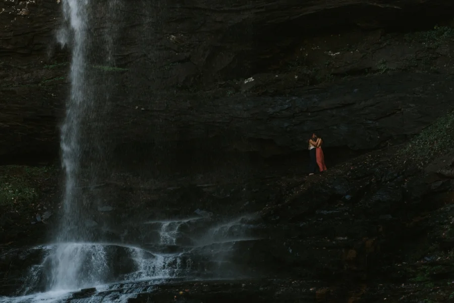 dark and romantic photo of a beautiful same sex couple in long maxi dresses cuddle and kiss by large waterfall at hemlock falls at cloudland canyon in northwest georgia