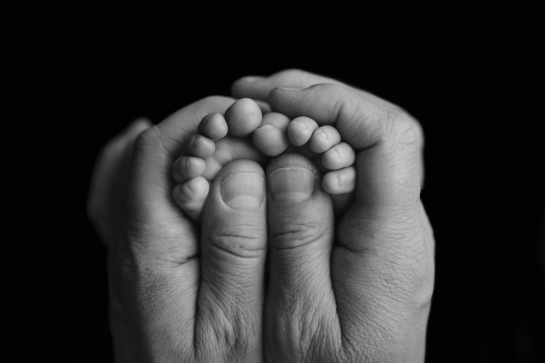 A black and white photograph a newborn baby's tiny feet with an adult's hands gently holding  The background is black, emphasizing the tiny feet, hands and special moment. Photo by Lenka Wills Photography, Bath UK