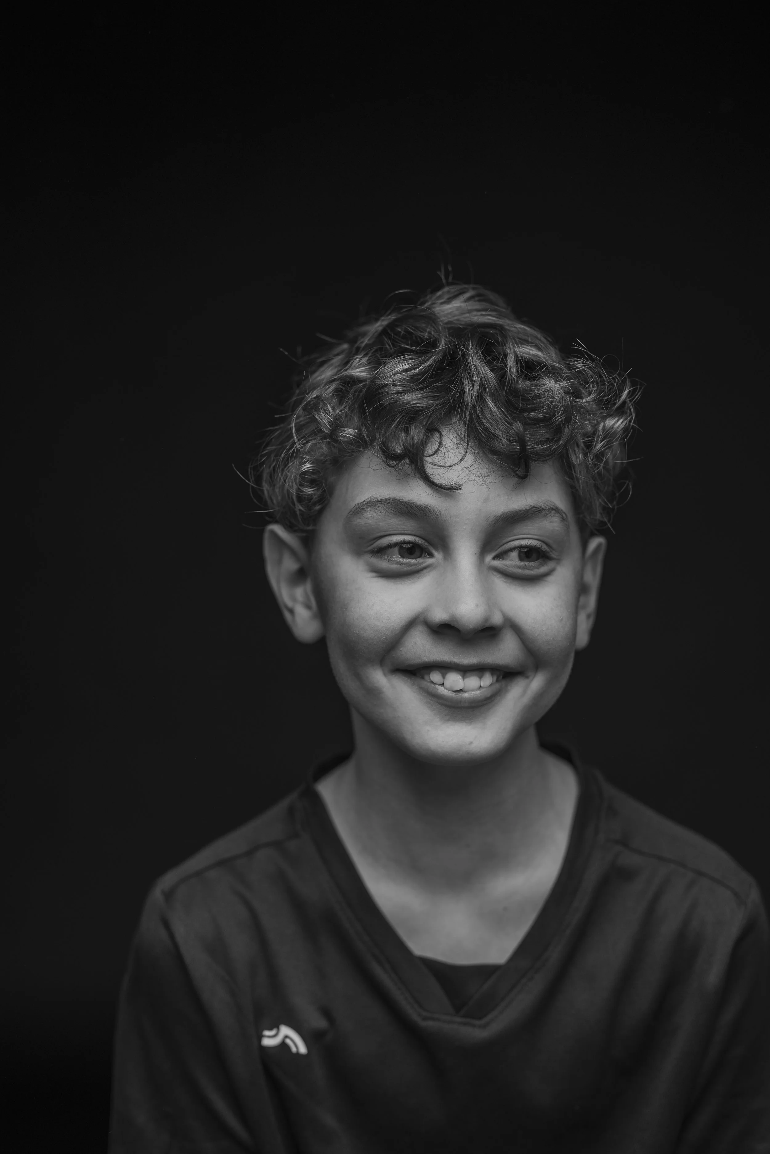 Black and white portrait of a smiling young boy with curly hair, against a dark background. Photo by Lenka Wills Photography, Bath UK