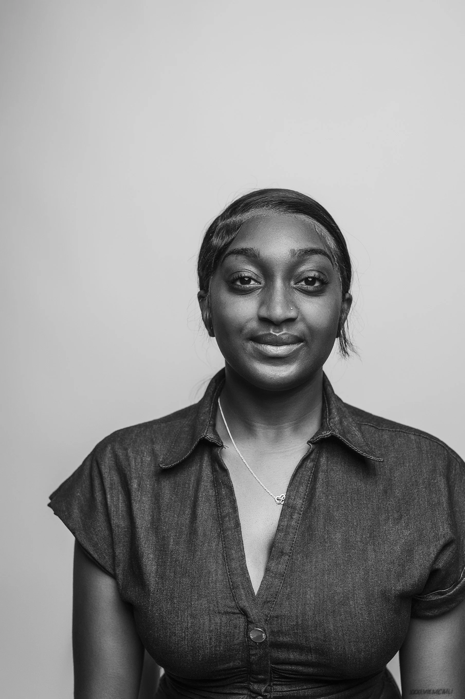 Close-up black and white portrait of a woman with dark hair tied back, wearing a denim shirt and a necklace, standing against a plain wall. Photo by Lenka Wills Photography, Bath UK.