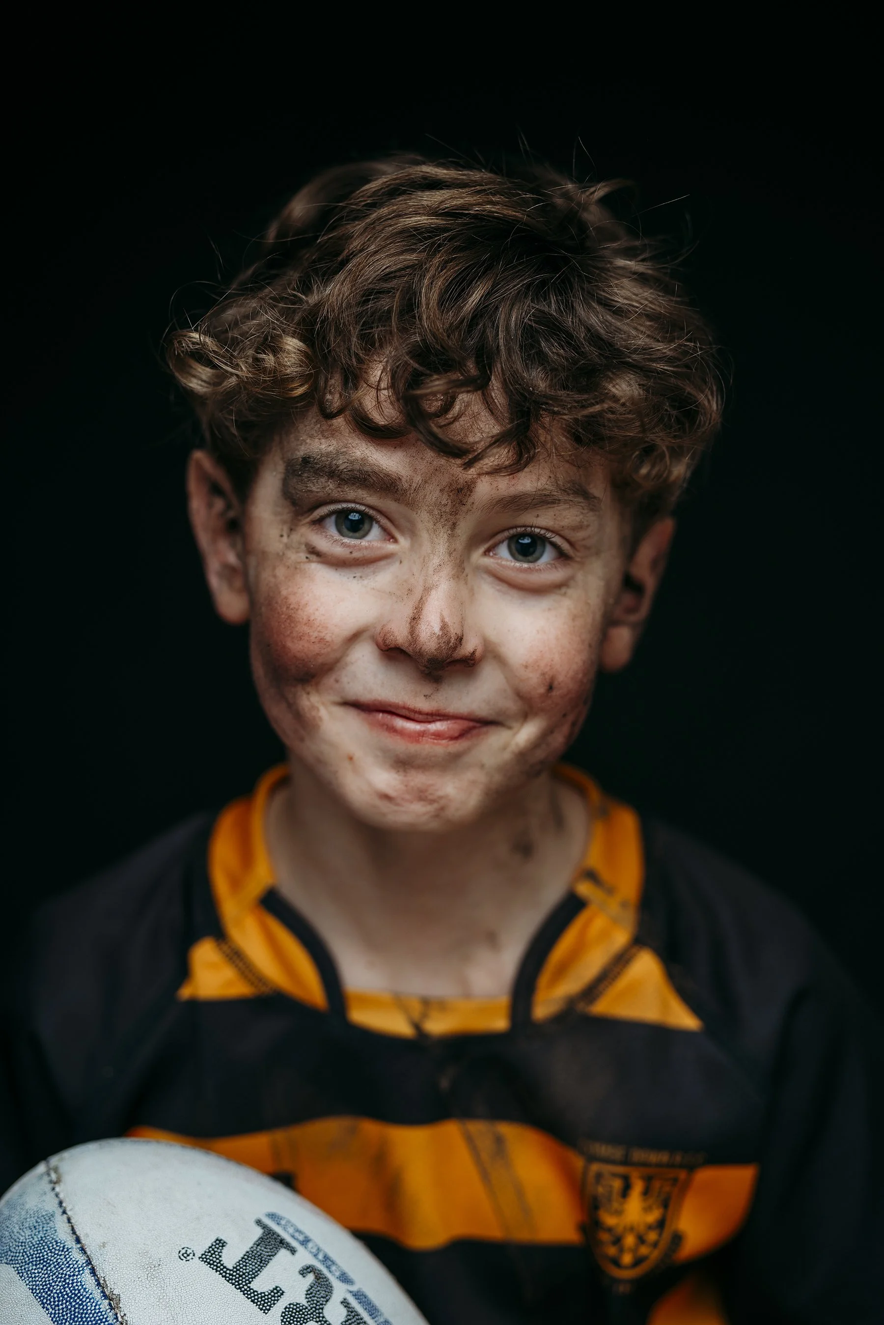 A young boy rugby player with curly brown hair, dirt on his face, and blue eyes smiling, wearing a black and orange rugby shirt, holding a rugby ball in front of a black background. Photo by Lenka Wills Photography, Bath UK