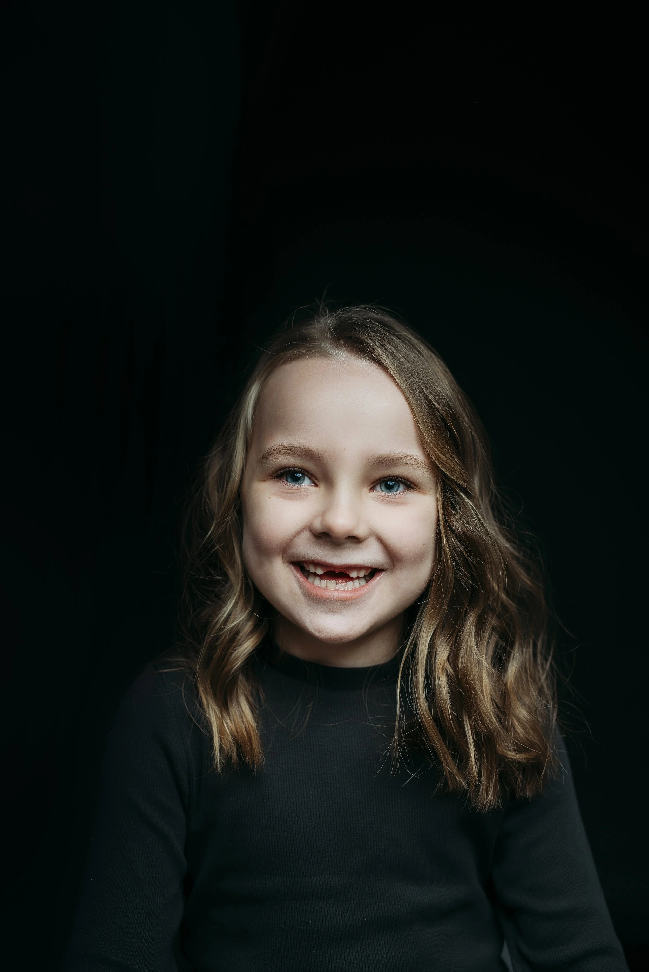 Young girl with long wavy hair smiling and looking up, wearing a dark top, against a black background. Photo by Lenka Wills Photography, Bath UK