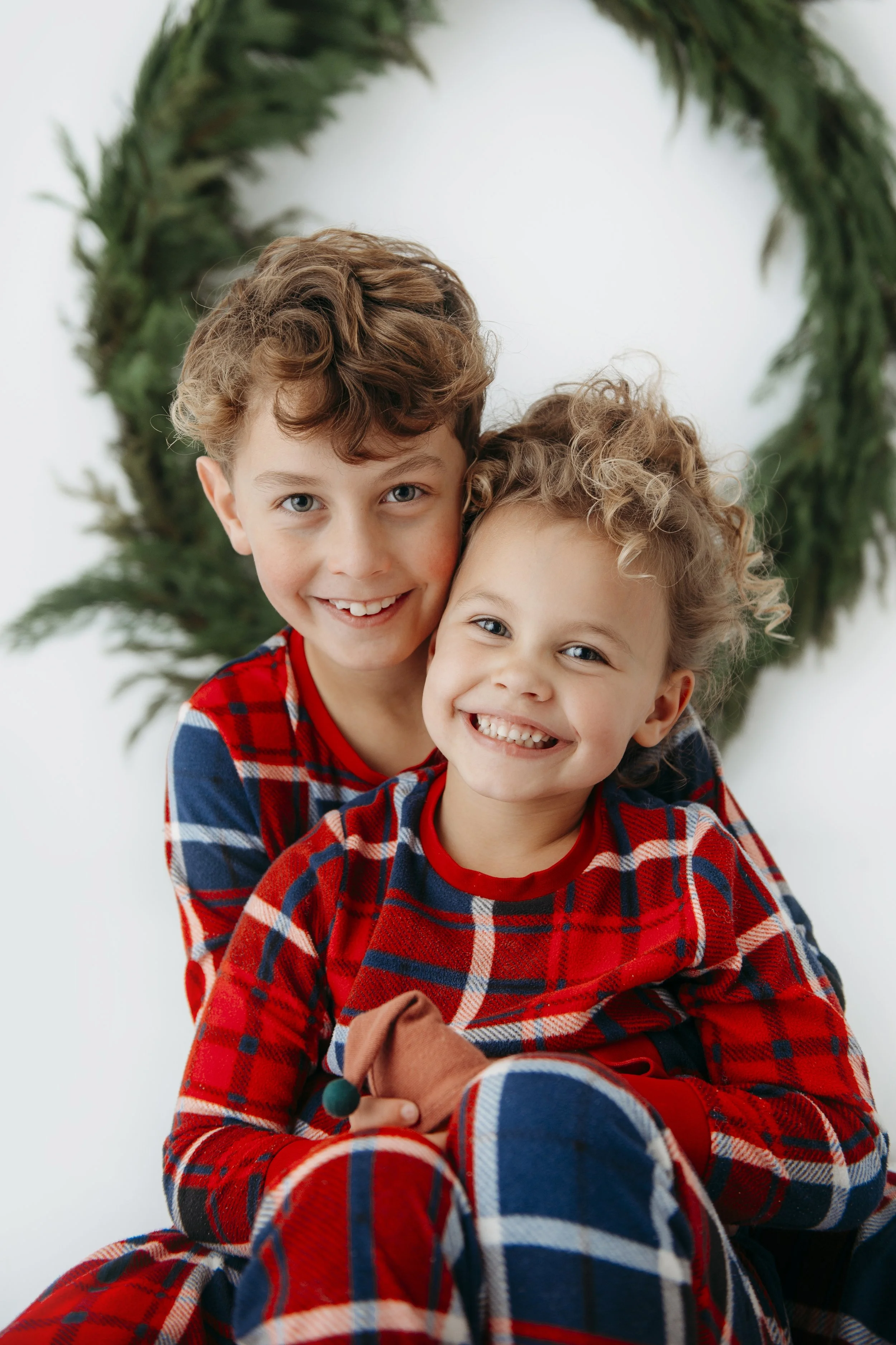 Two smiling young children wearing red and blue plaid pajamas, sitting together in front of a white background with a green wreath for a christmas photo shoot by Lenka Wills Photography, Bath UK.
