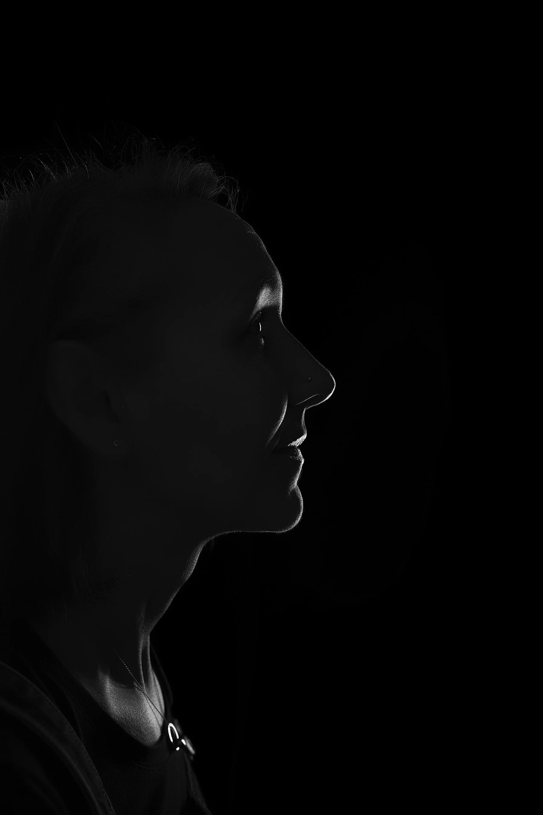 Side profile of a woman's face in dramatic lighting, showing details of hair, nose, lips, and chin, against a black background. Photo by Lenka Wills Photography, Bath UK