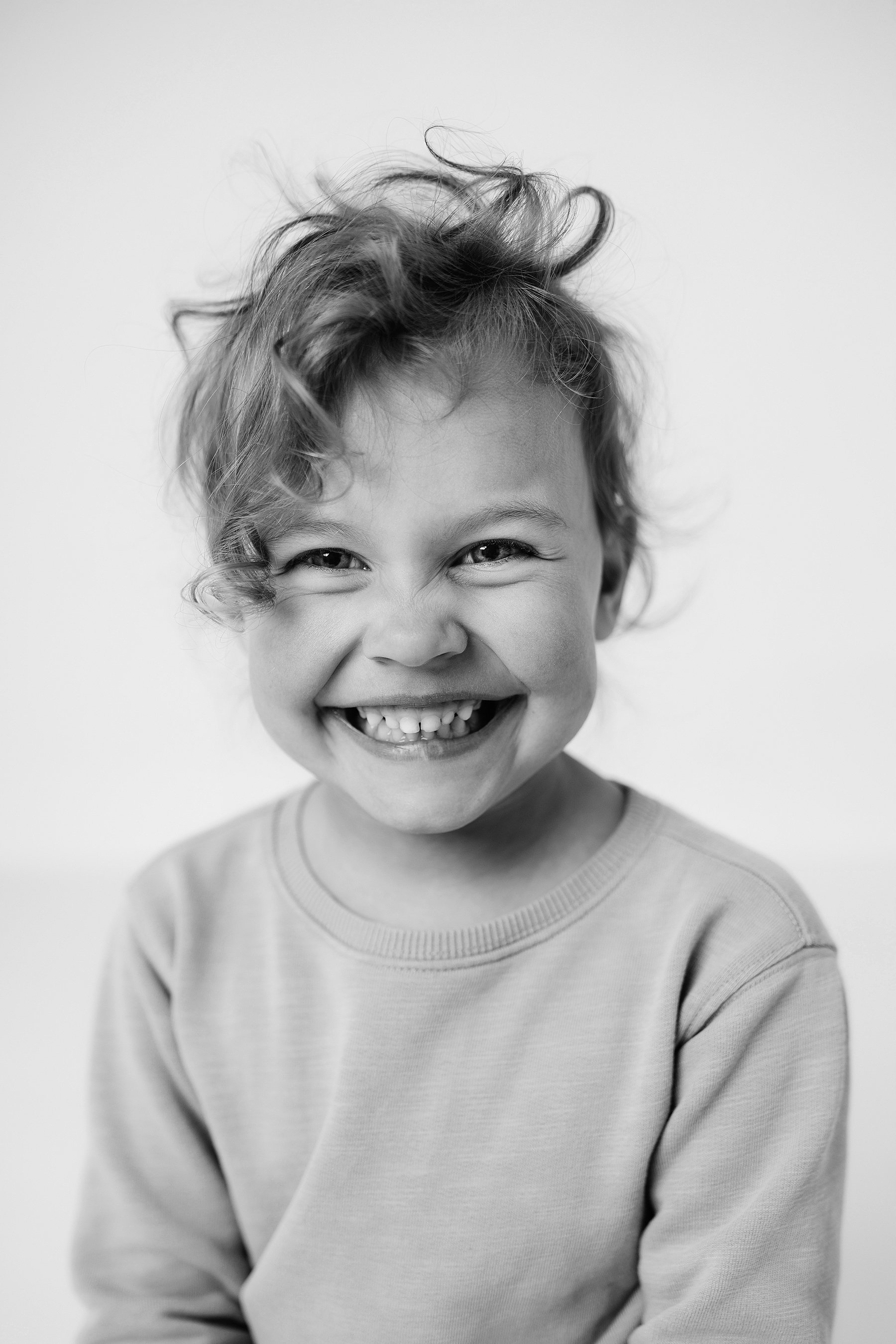 A smiling child with curly hair is looking at the camera, wearing a light-colored sweatshirt, against a plain background, photo by Lenka Wills Photography, Bath UK