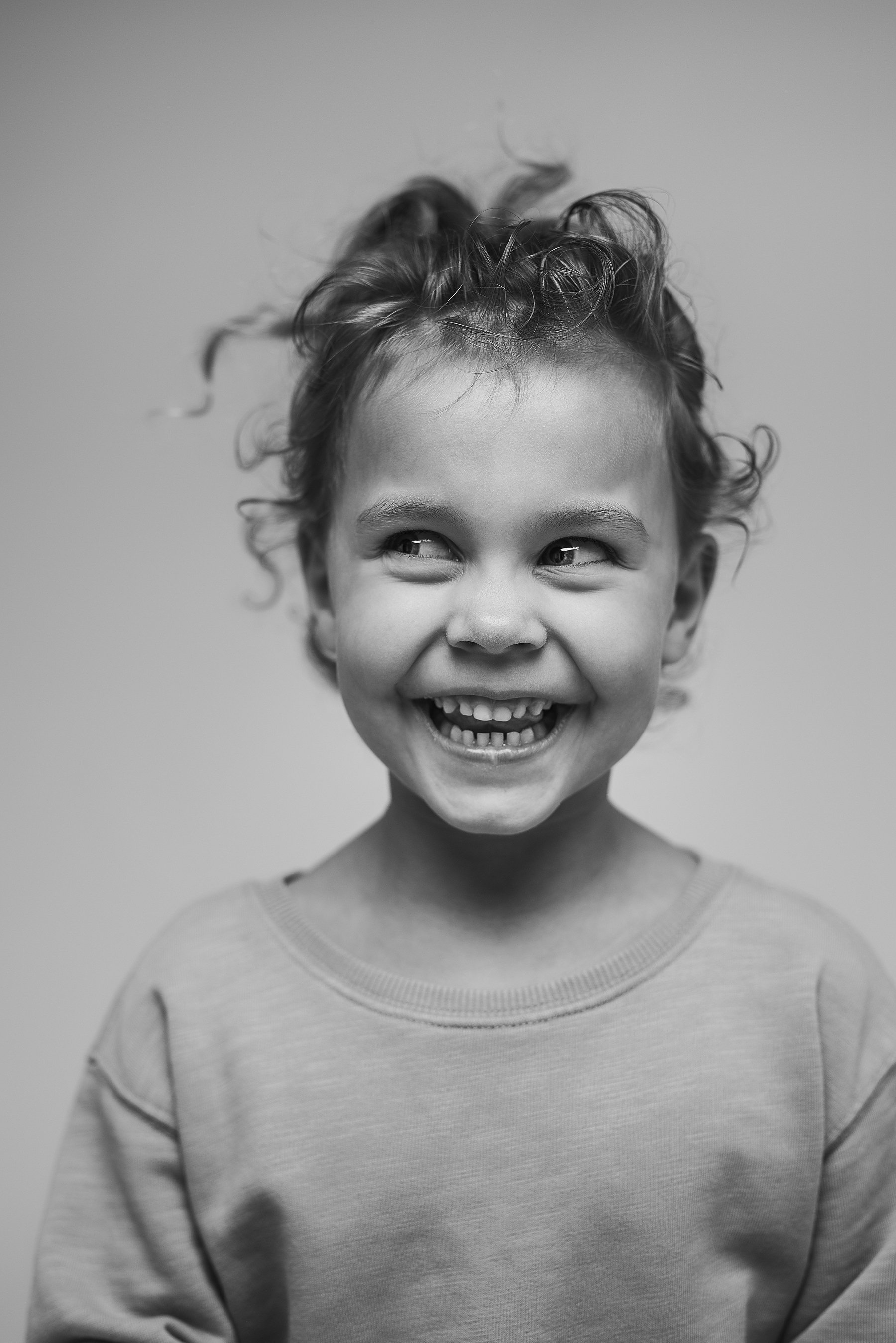 A smiling young child with curly hair wearing a sweatshirt, captured in black and white, looking slightly to the side. Photo by Lenka Wills Photography, Bath UK