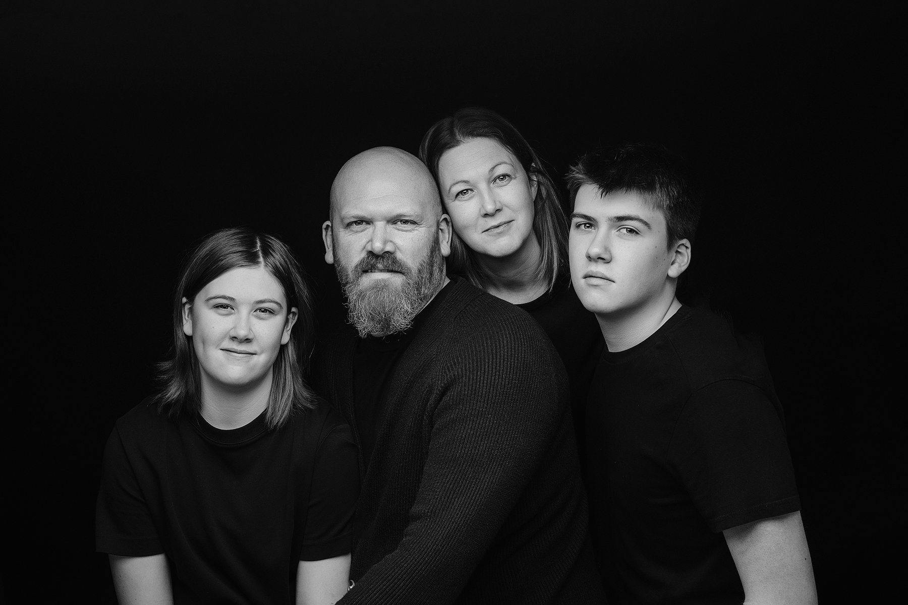Black and white portrait of a family of four, including dad, mum and their teenage children, against a dark background. Photo by Lenka Wills Photography, Bath UK