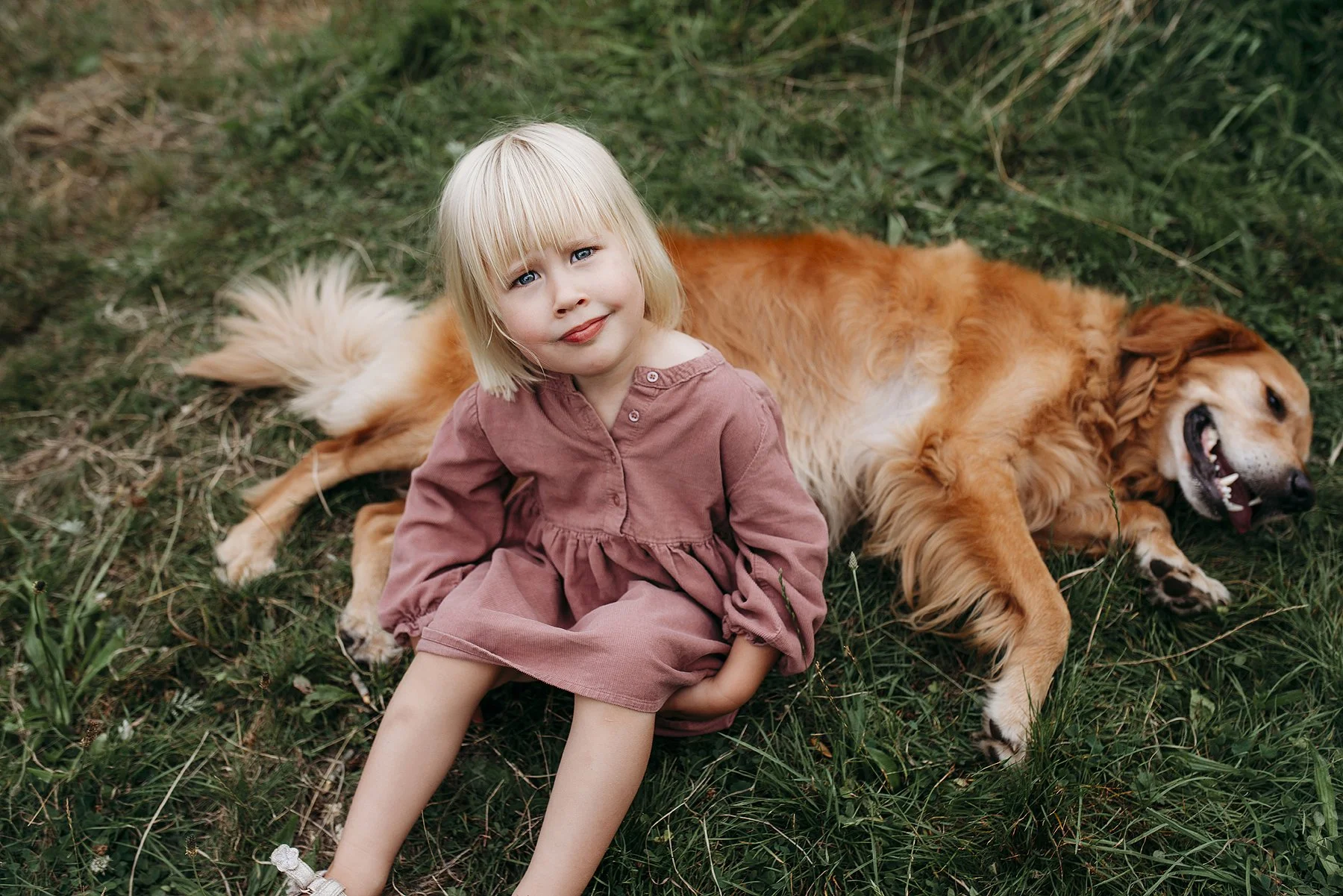 A young girl with blonde hair, blue eyes and a pink dress  sitting on grass next to a golden retriever that is lying on its side with its mouth open, in an outdoor setting. Photo by Lenka Wills Photography, Bath UK