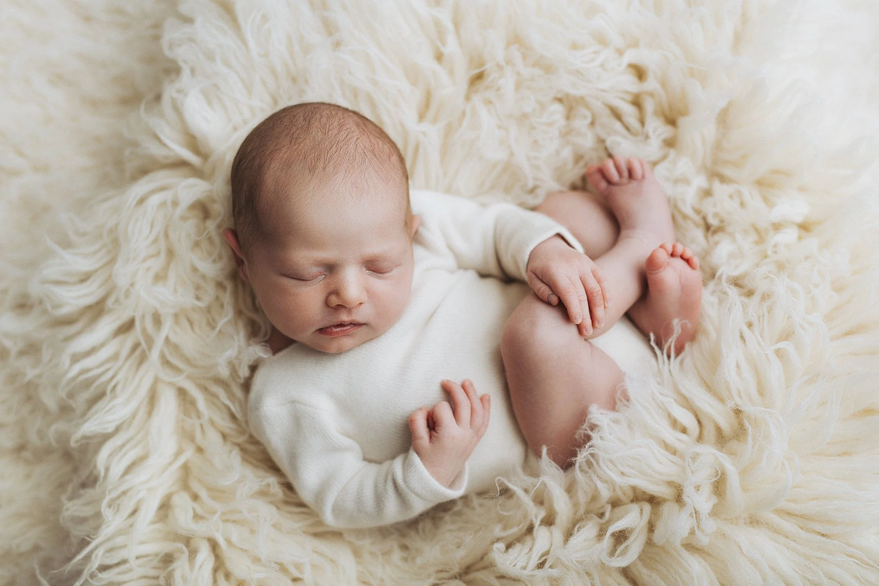 A sleeping newborn baby lying on a fluffy woolly blanket, wearing a white babygrow, with one hand resting on their chest and the other on their leg. Photo by Lenka Wills Photography, Bath UK