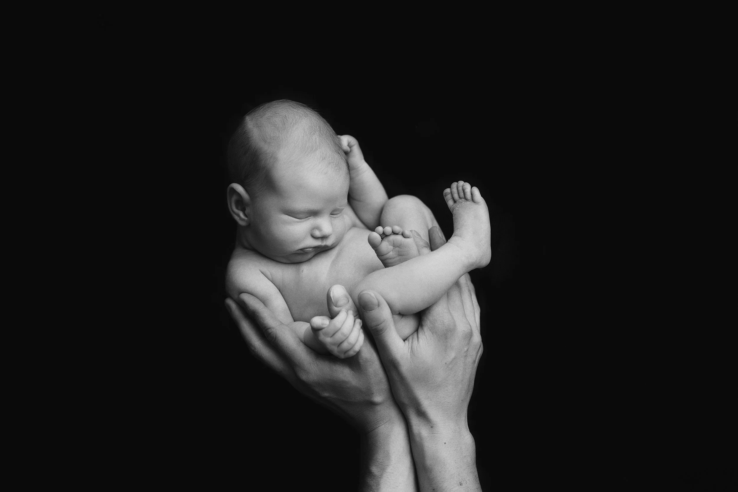 Black and white photo of a tiny newborn baby cradled in an adult's hand, with the baby's head resting on the adult's fingers and an arm wrapped around the baby's body. Photo by Lenka Wills Photography, Bath UK