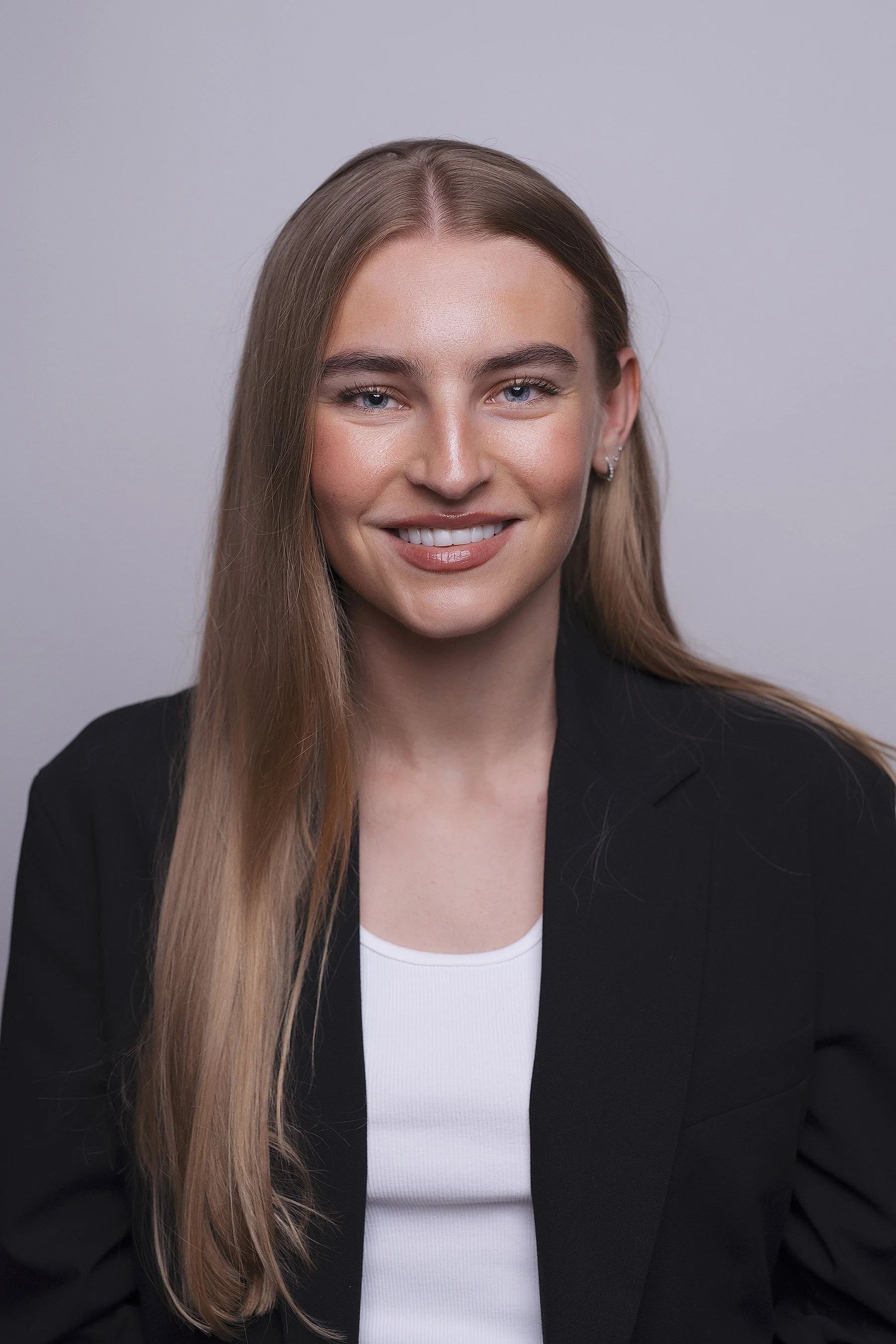 A young woman with long, straight, light brown hair, blue eyes, and a friendly smile, wearing a black blazer over a white top, against a plain gray background. Corporate portrait photography by Lenka Wills, Bath UK.