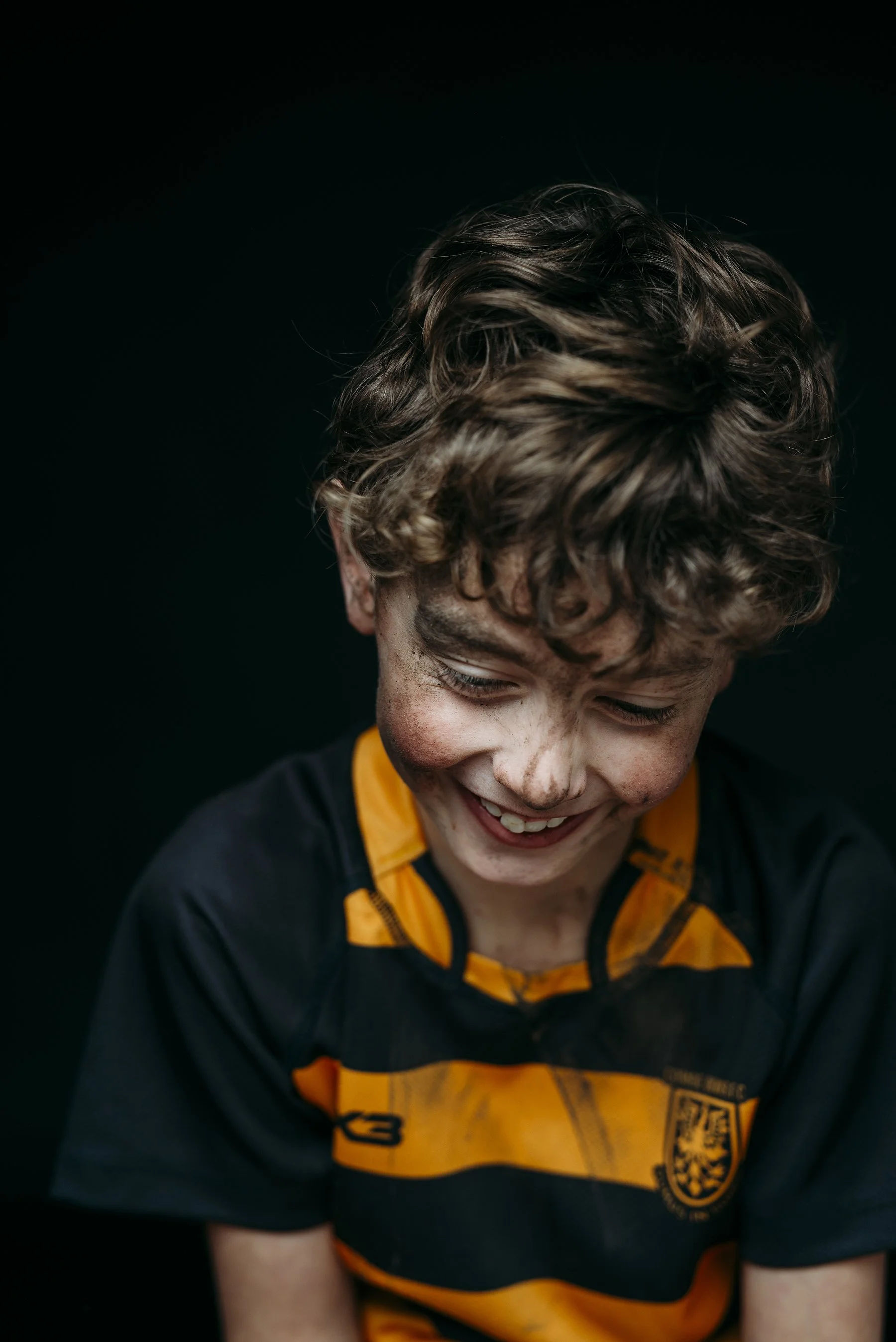 A boy with curly hair smiling, wearing a black and orange rugby shirt, against a black background. Photo by Lenka Wills Photography, Bath UK