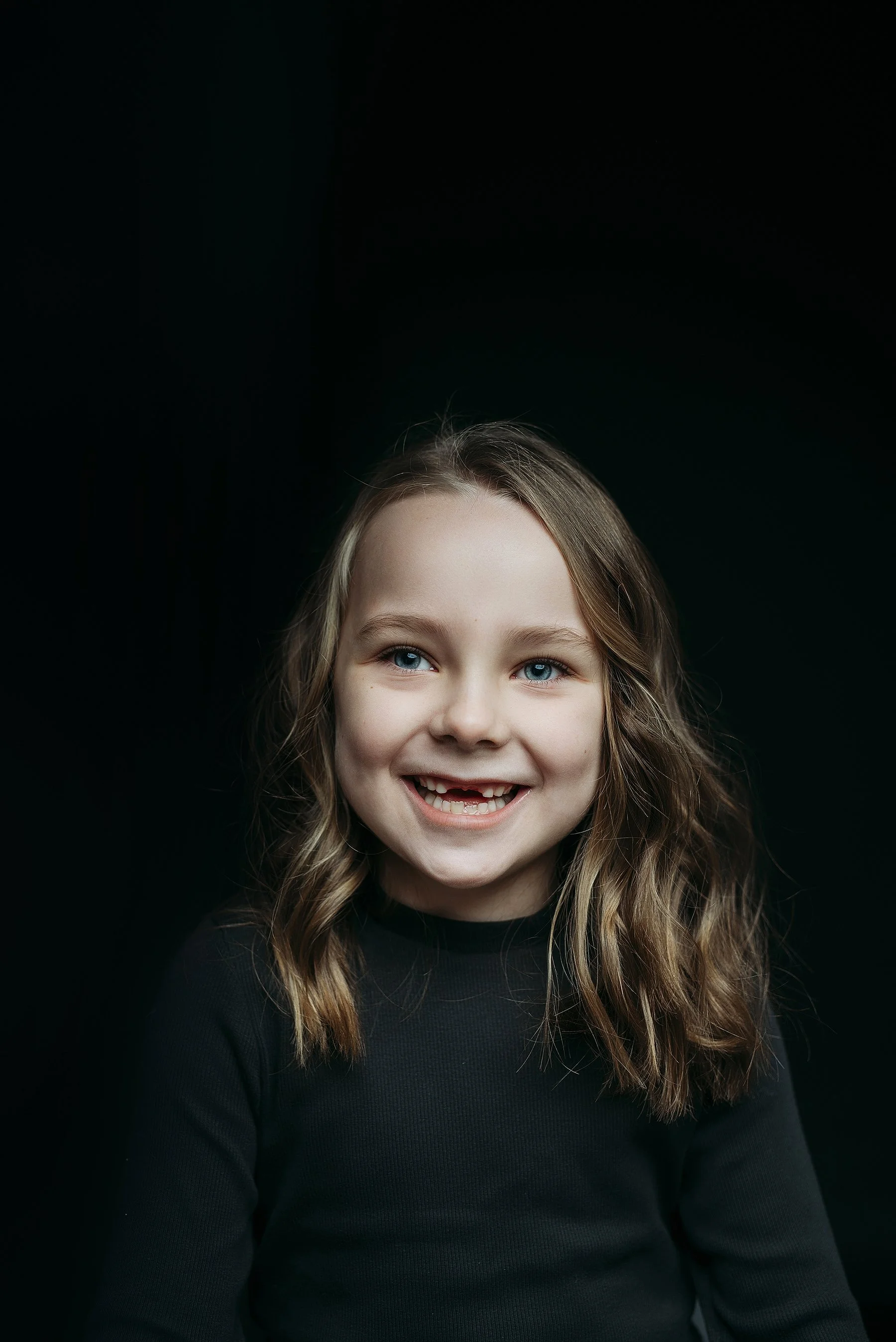 Close-up of a smiling young girl with blue eyes and wavy brown hair, wearing a black shirt, set against a black background. Photo by Lenka Wills Photography, Bath UK