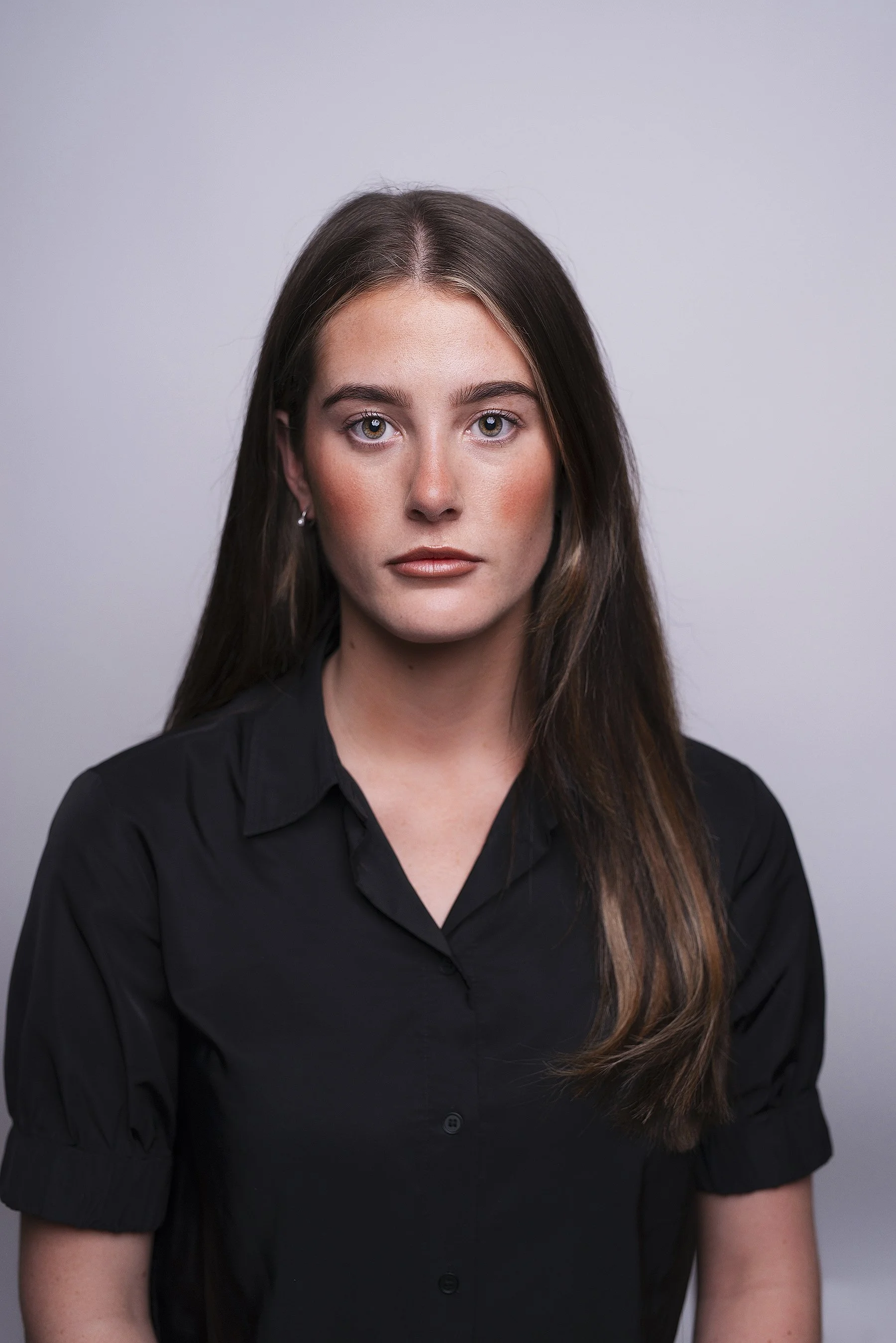Portrait of a young woman with long brown hair, wearing a black button-up shirt, standing against a plain gray background. Corporate portrait photography by Lenka Wills, Bath UK.