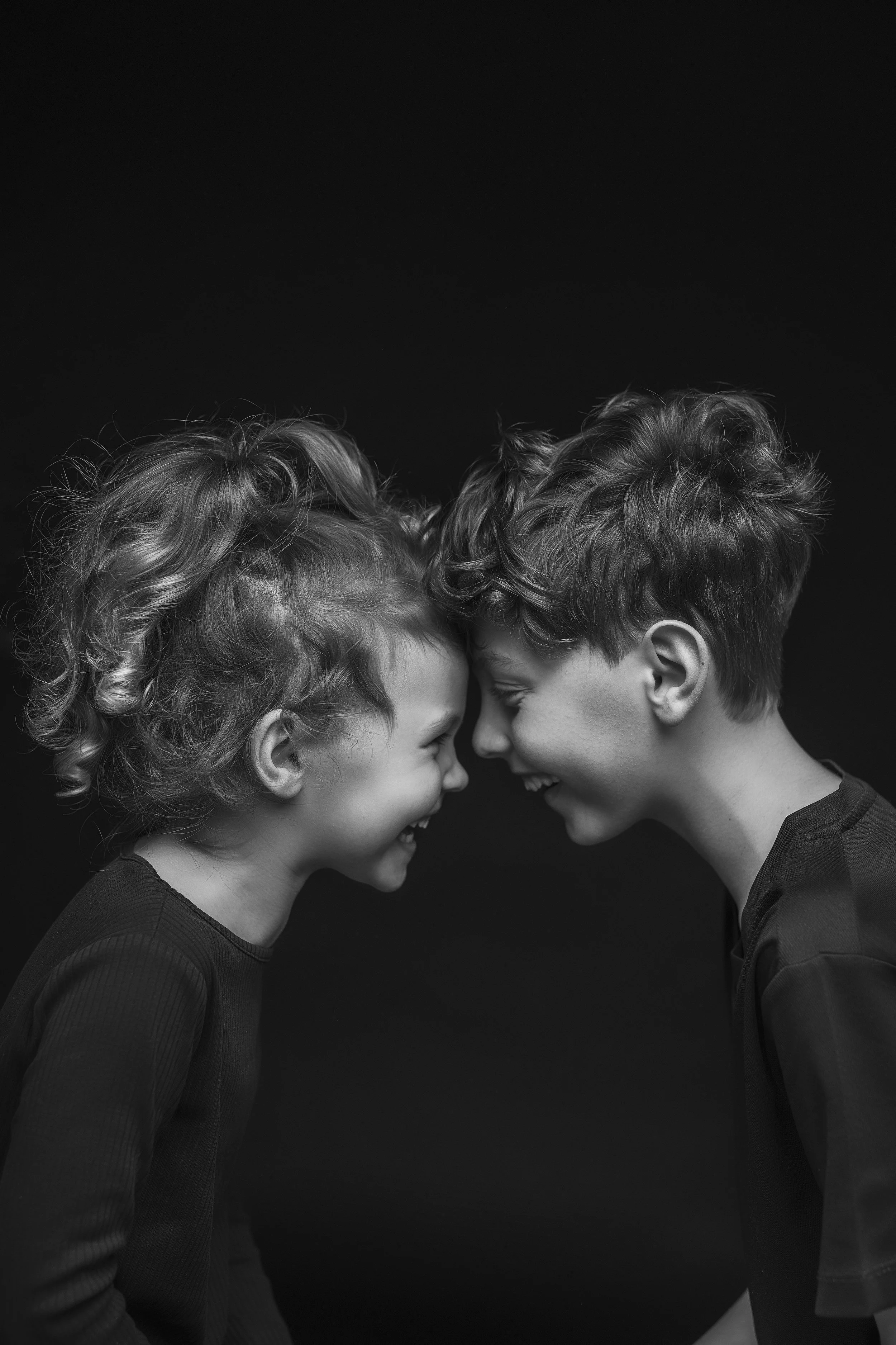 A black and white photo of two children, a girl with curly hair and a boy with short, wavy hair, touching foreheads and smiling at each other against a black background. Photo by Lenka Wills Photography, Bath UK
