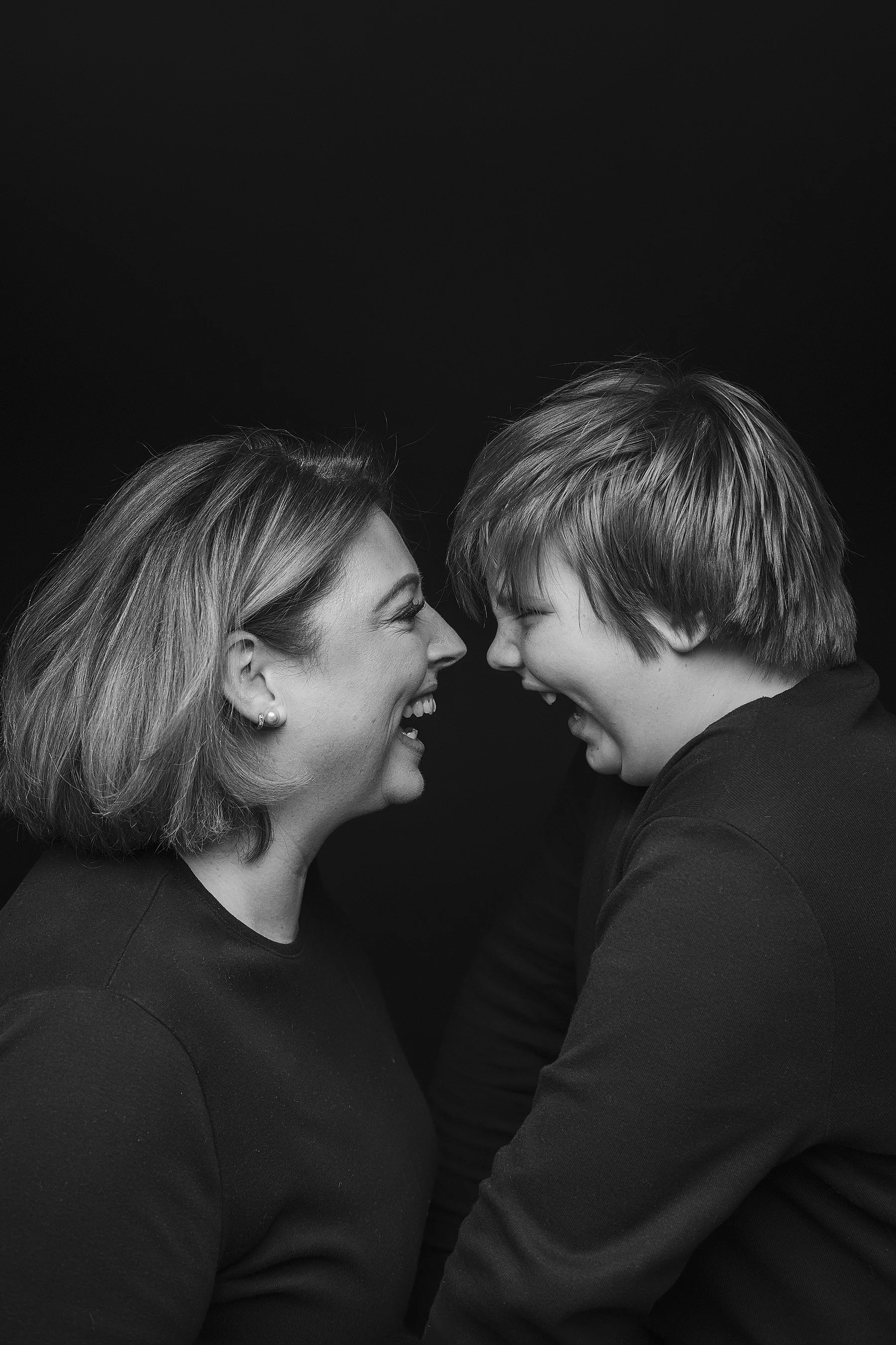 A black and white photo of a mum and child facing each other, smiling, with their foreheads touching, close together, against a black background. Photo by Lenka Wills Photography, Bath UK