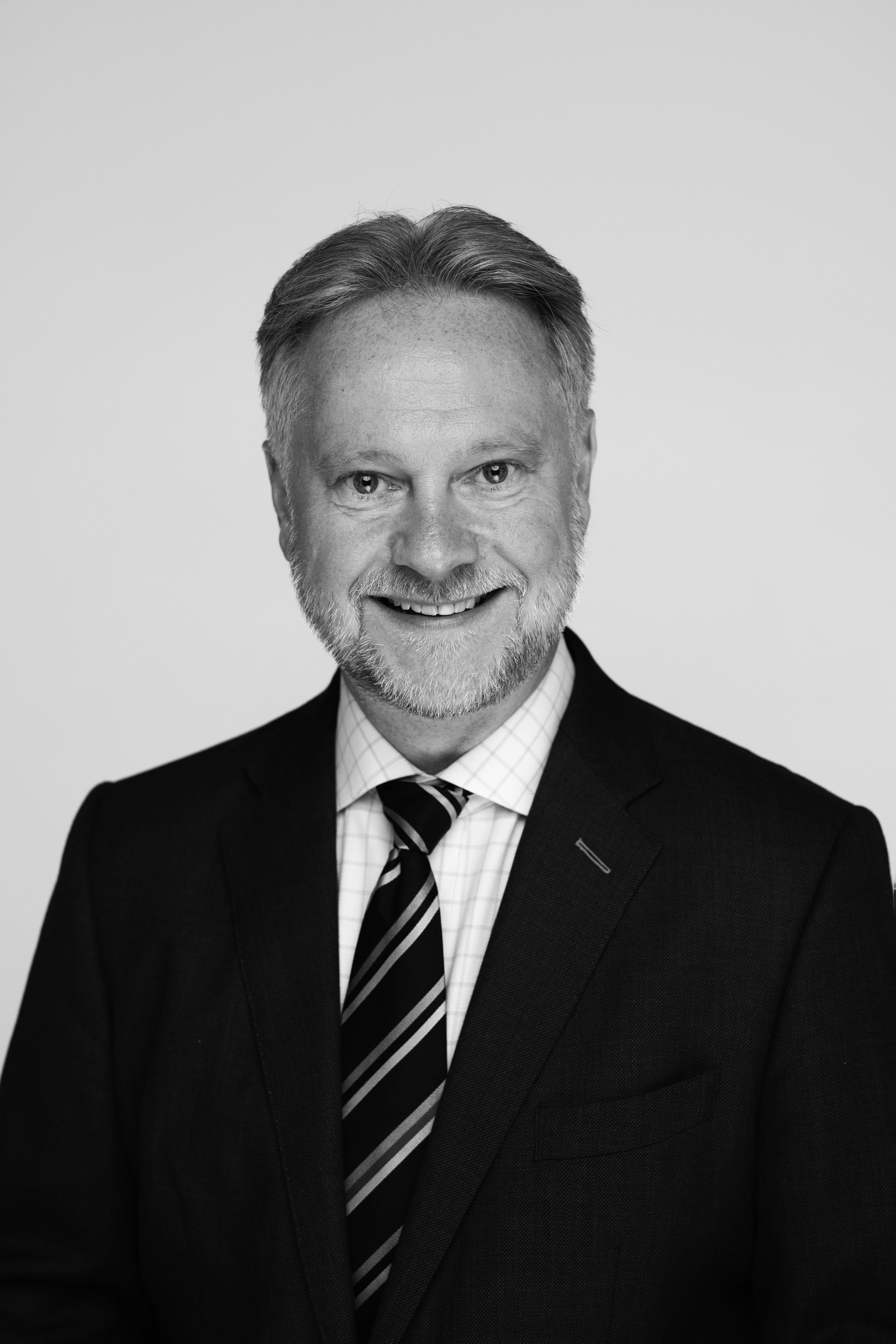 Headshot of a middle-aged man with gray hair and beard, wearing a dark suit, patterned dress shirt, and striped tie, smiling against a plain background.