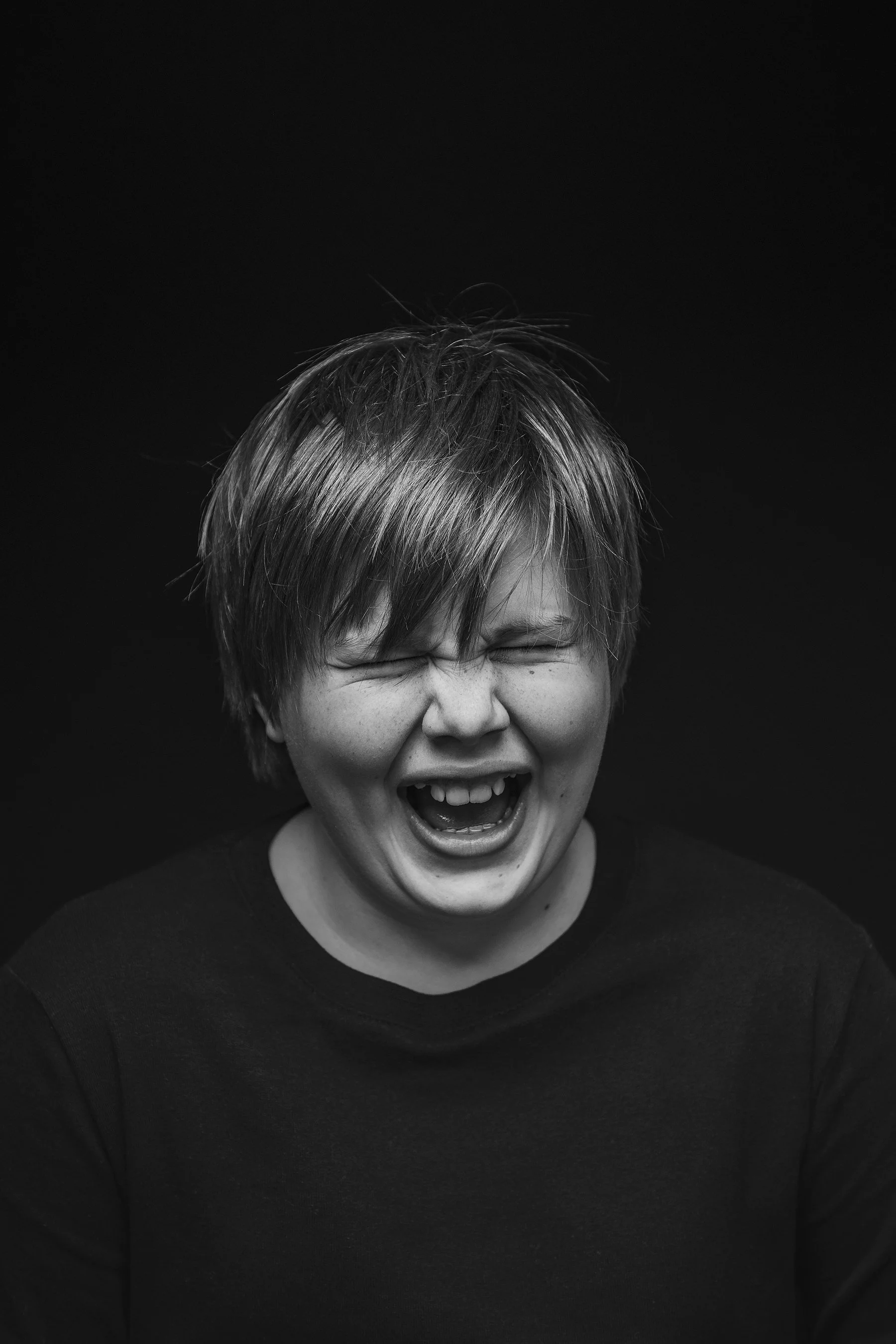 A boy with hair over his face laughing with eyes closed, wearing a black shirt, against a black background. Photo by Lenka Wills Photography, Bath UK