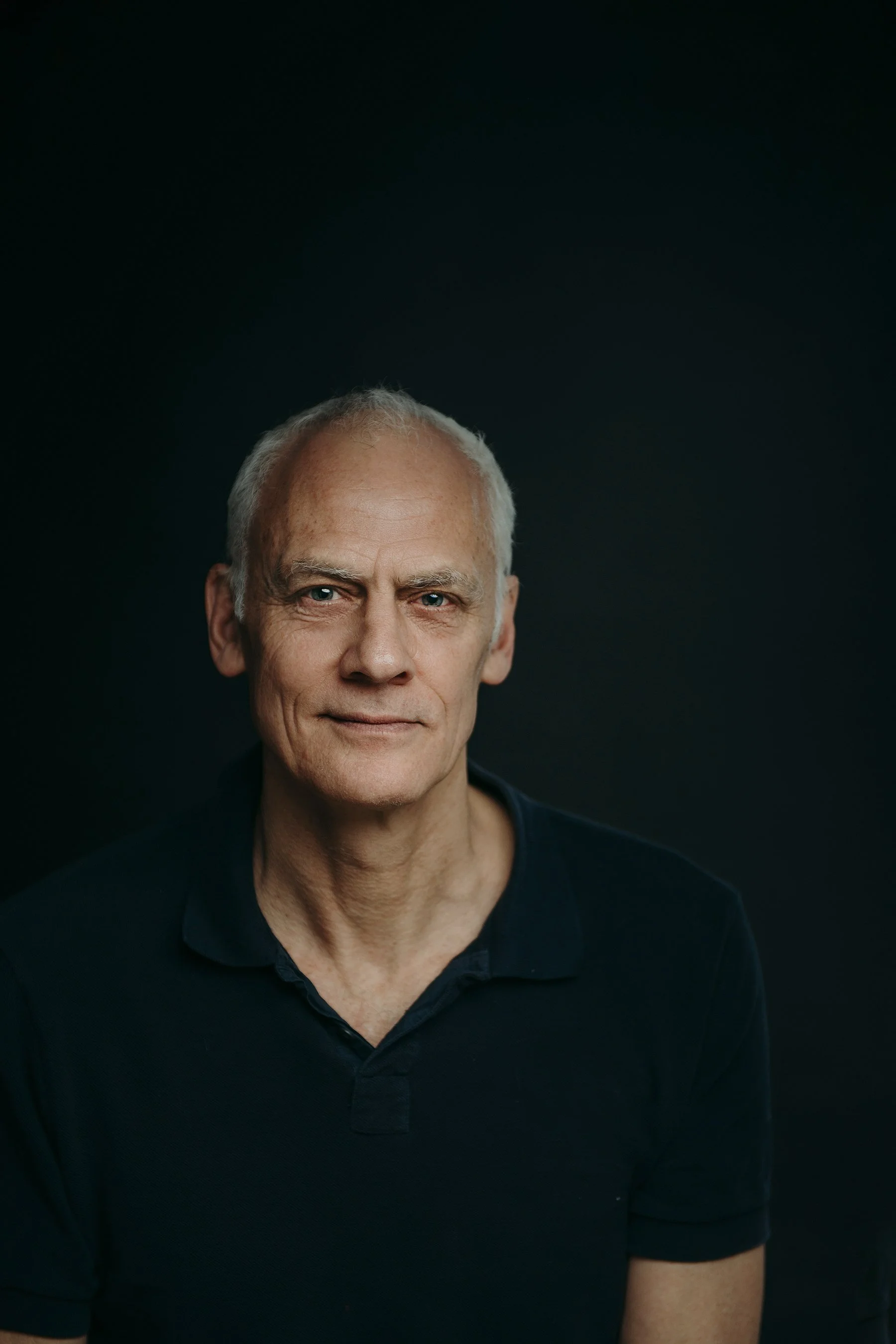 A portrait of an older man with short gray hair, blue eyes, and fair skin, wearing a dark blue shirt, against a black background. Photo by Lenka Wills Photography, Bath UK