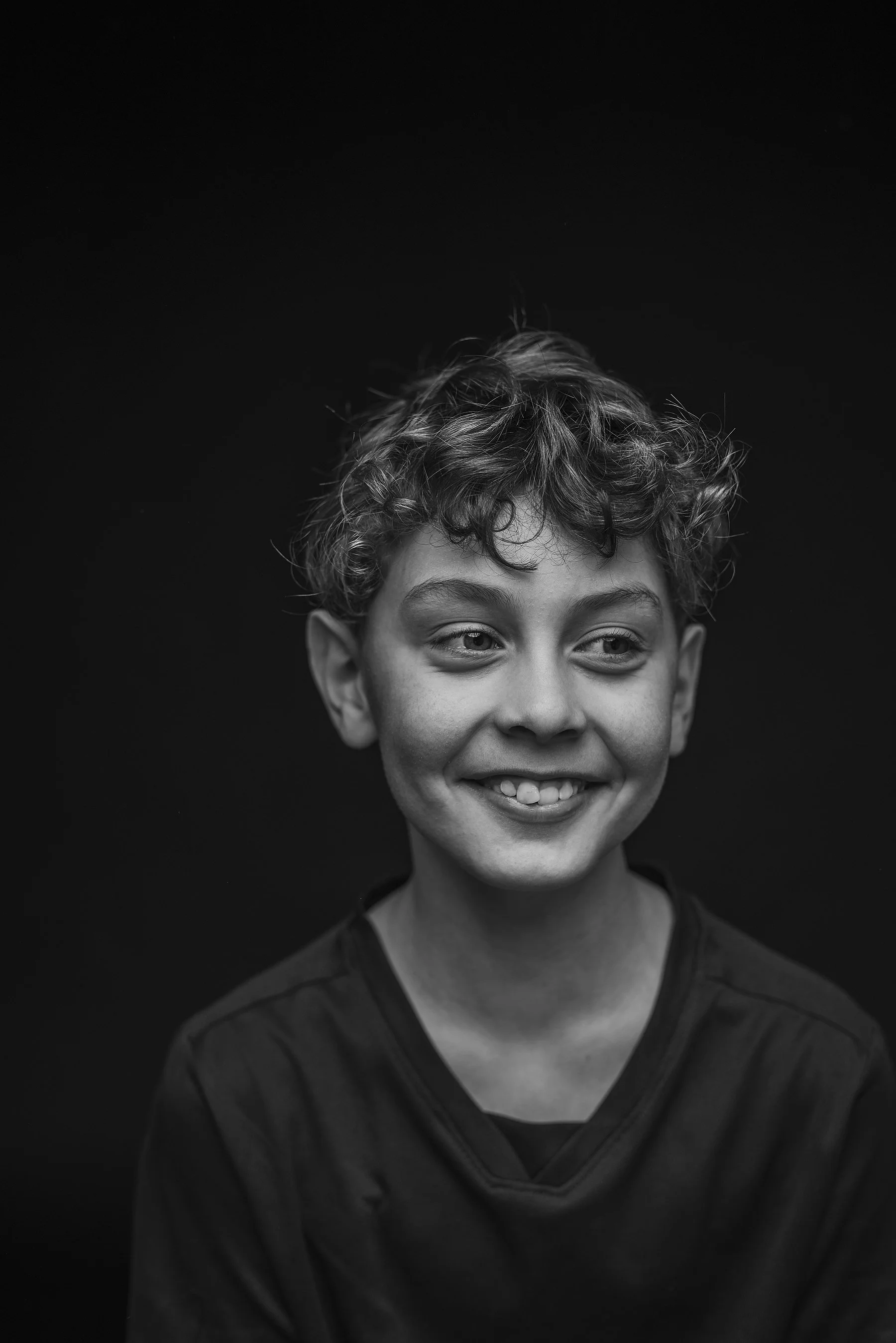 Black and white photo of a young boy with curly short hair smiling, wearing a dark shirt, against a black background. Photo by Lenka Wills Photography, Bath UK