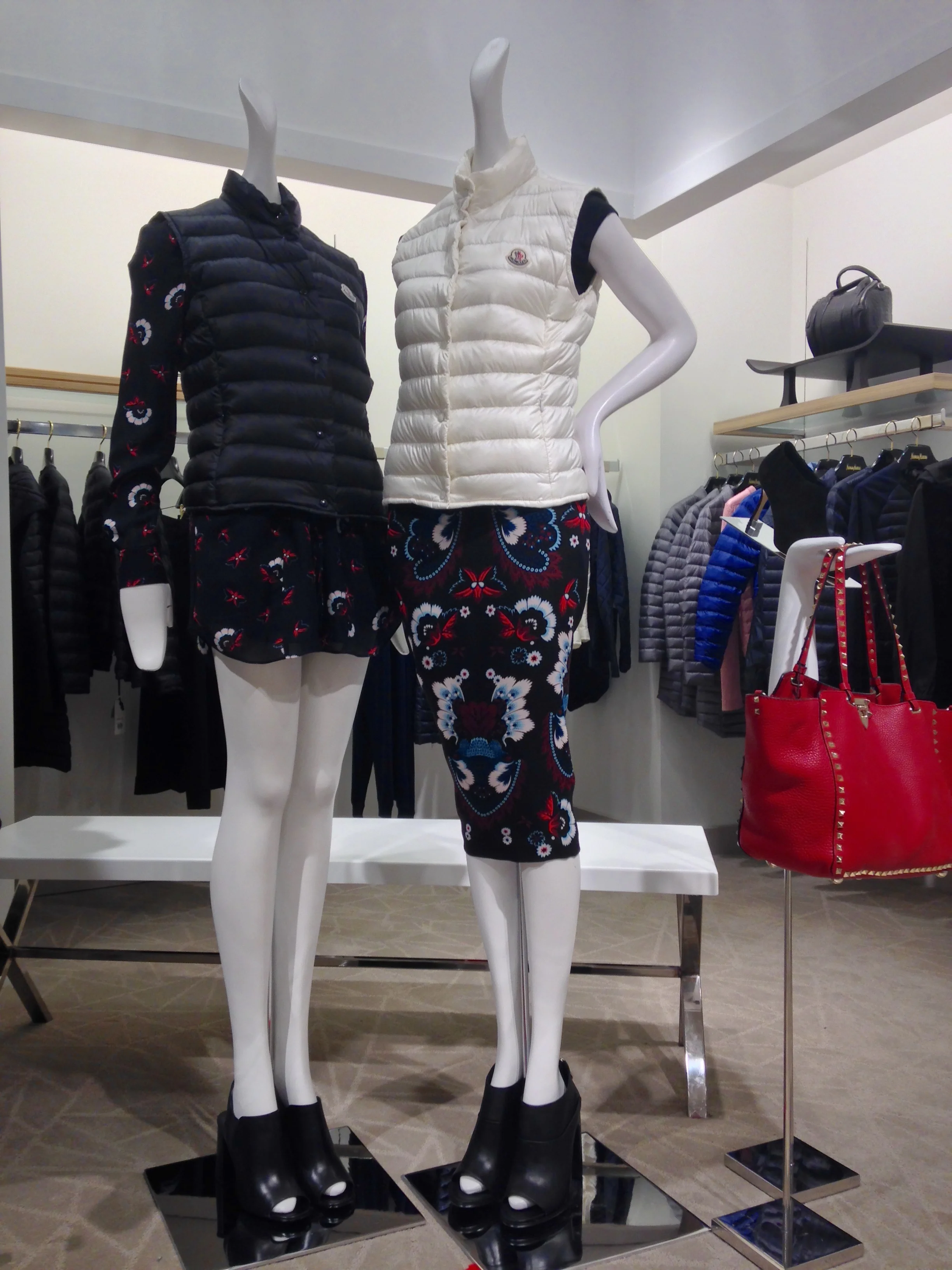 Two mannequins dressed in puffy vests and floral skirts, one in black and one in white, displayed in a clothing store with racks of apparel and a red handbag visible in the background.