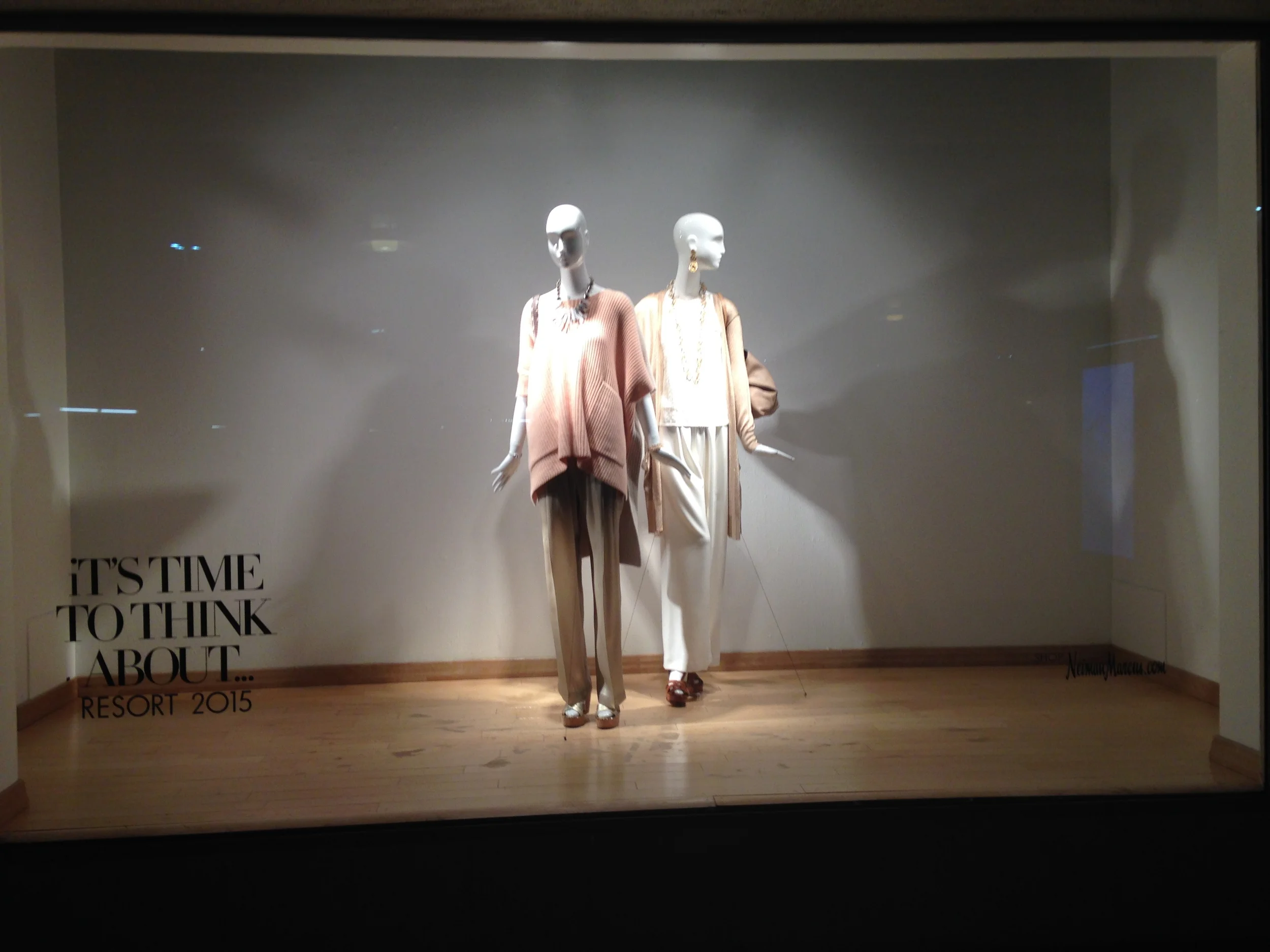 Two mannequins dressed in beige and brown clothing, with handbags and jewelry, are displayed in a store window with the text 'It's time to think about... Resort 2015'.