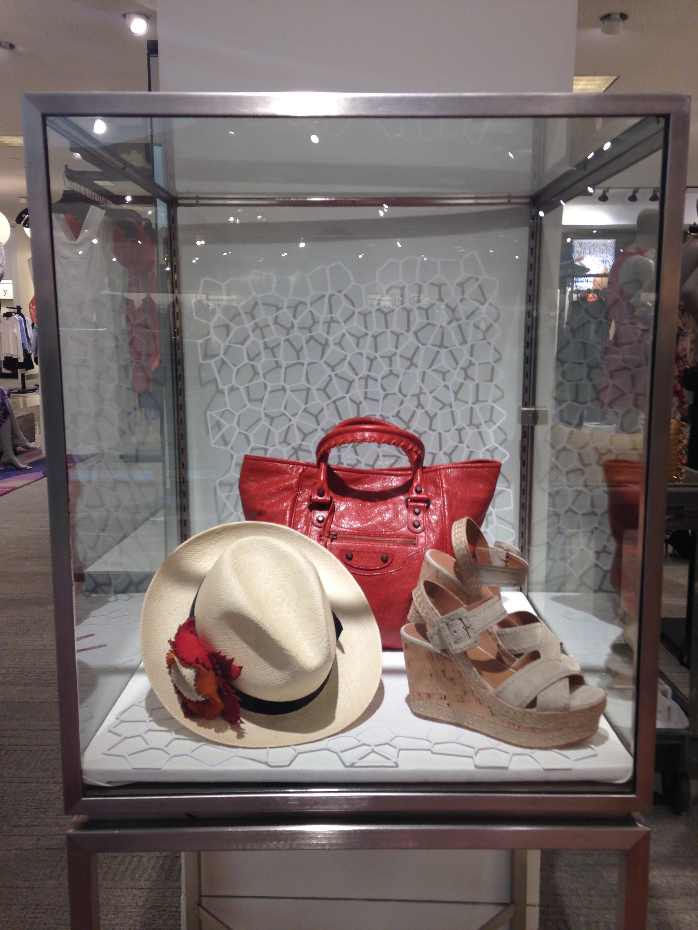Display case with a red handbag, straw hat with floral decoration, beige sandals, and other accessories.