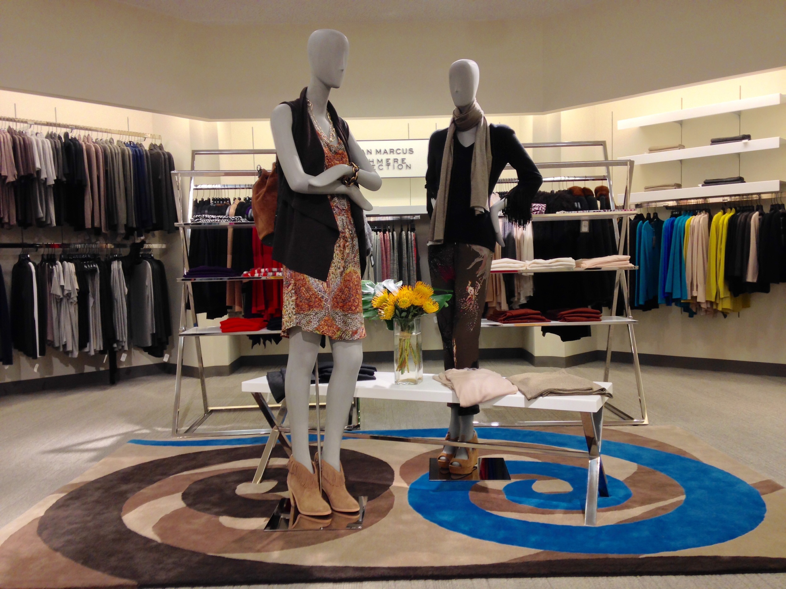 Mannequin wearing a patterned dress, gray blazer, and scarf, holding a handbag, and another mannequin wearing a black jacket and floral top, with clothing racks and shelves in the background.