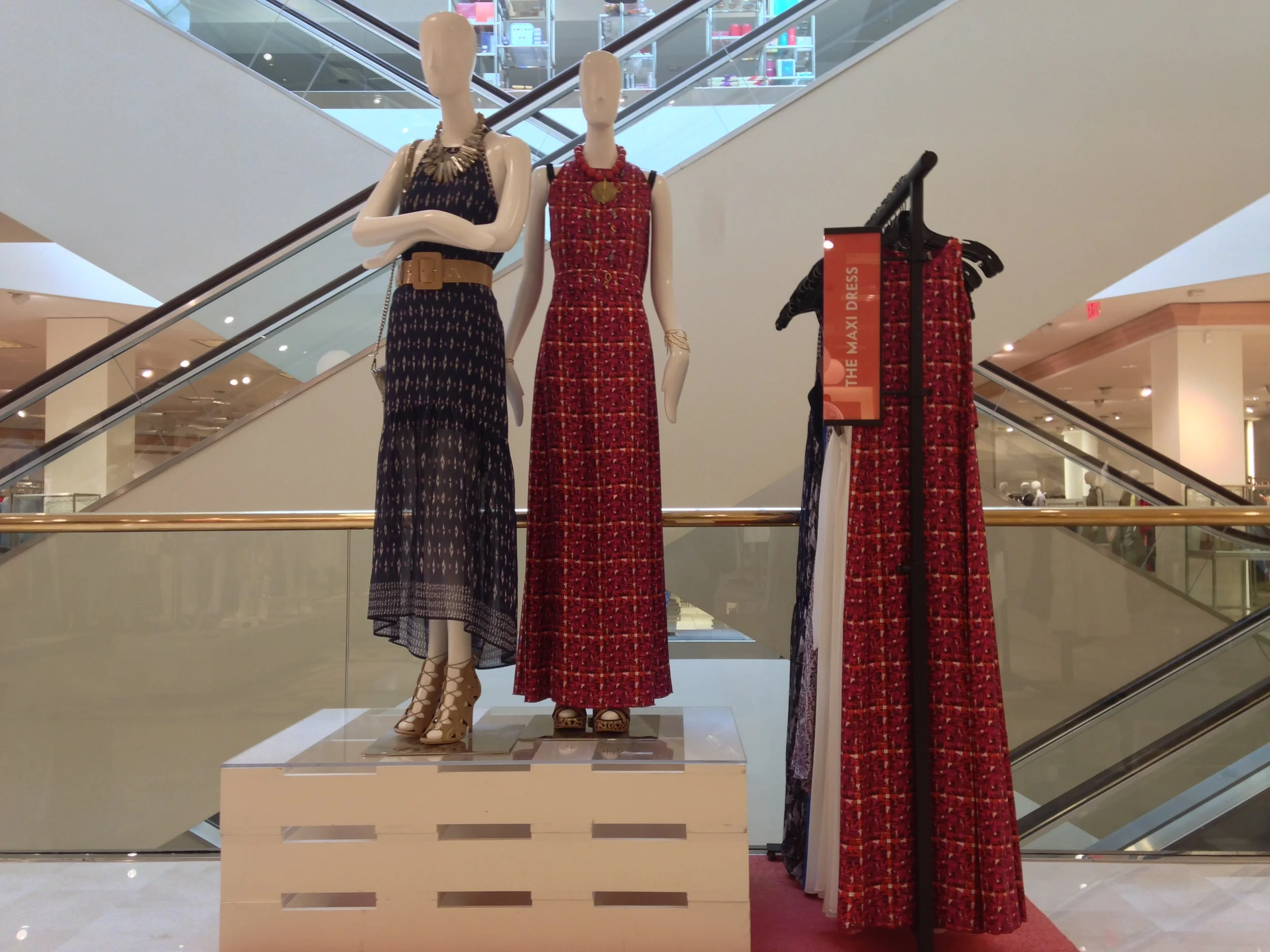 Display of mannequins wearing colorful dresses in a shopping mall with a staircase in the background.