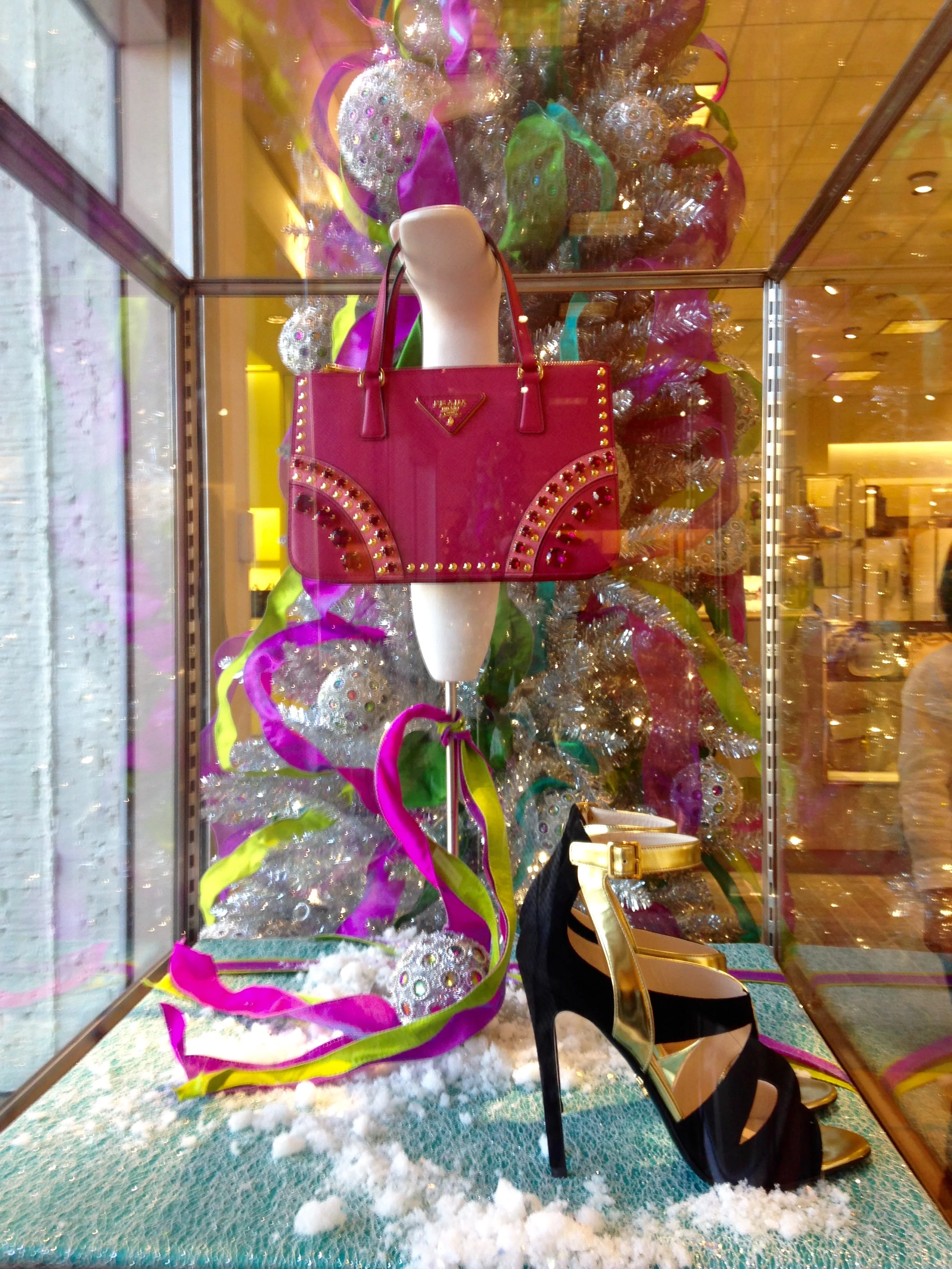 Display window with colorful designer handbags, including a prominent red and black Prada bag, surrounded by decorative ribbons, ornaments, and faux snow, in a shopping mall.