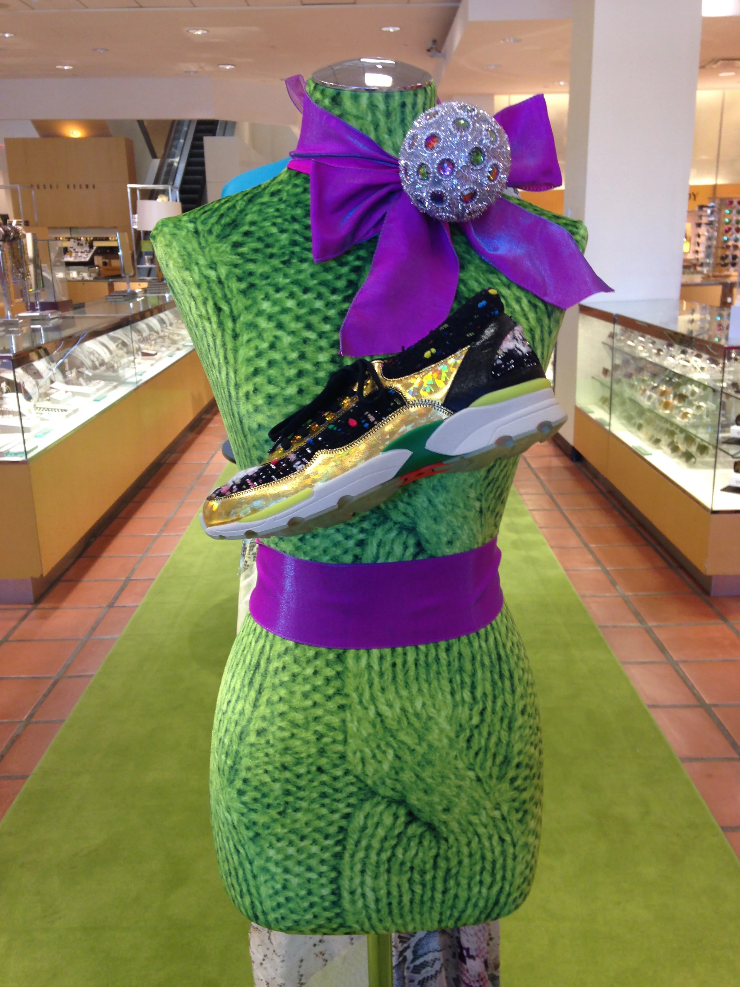 A mannequin torso decorated with a green textured fabric, a large purple ribbon, a rhinestone-encrusted decorative orb, and a multicolored sneaker.