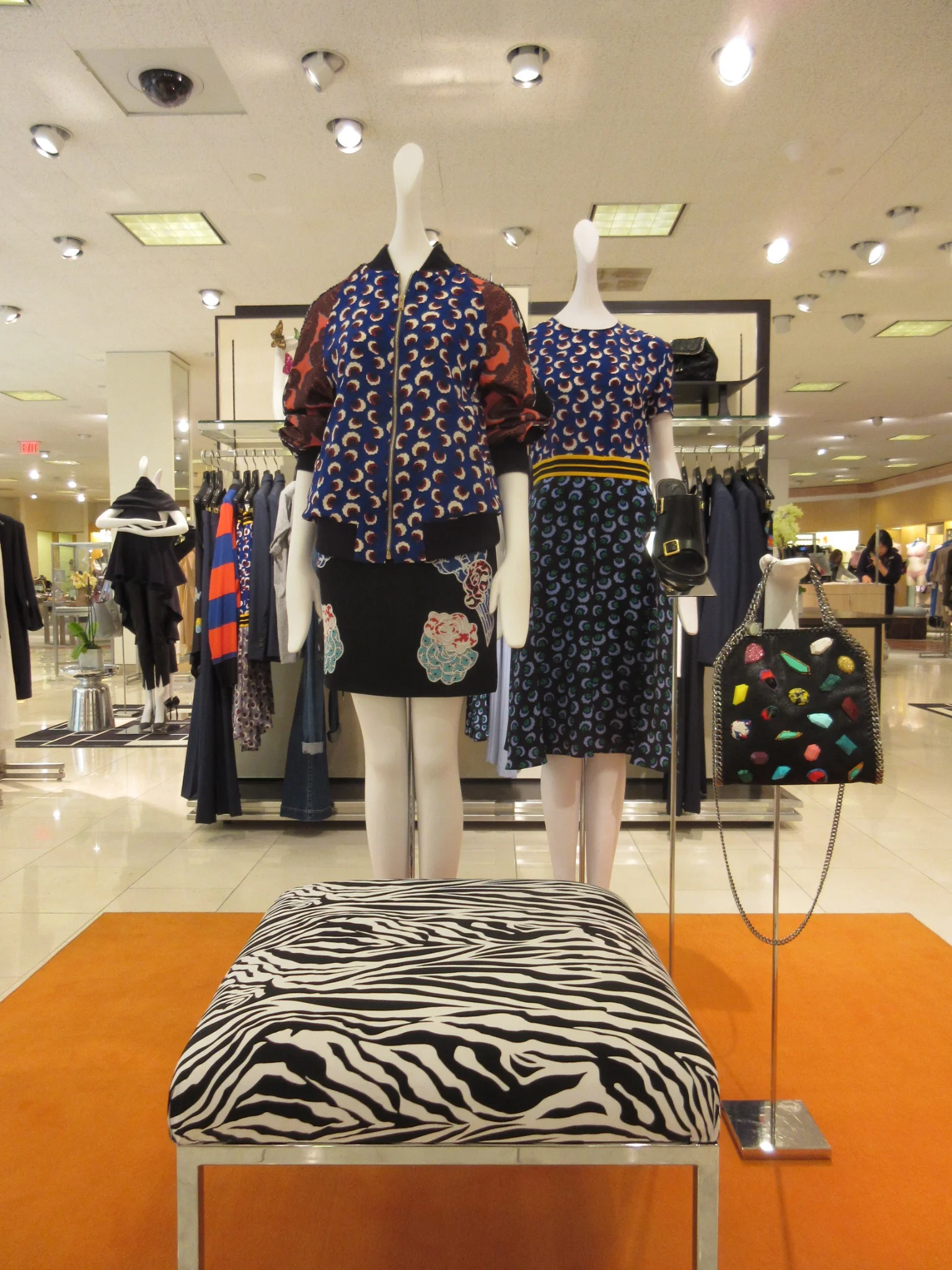 Two mannequins dressed in colorful patterned clothing stand on display in a clothing store, with clothing racks and shelves in the background.